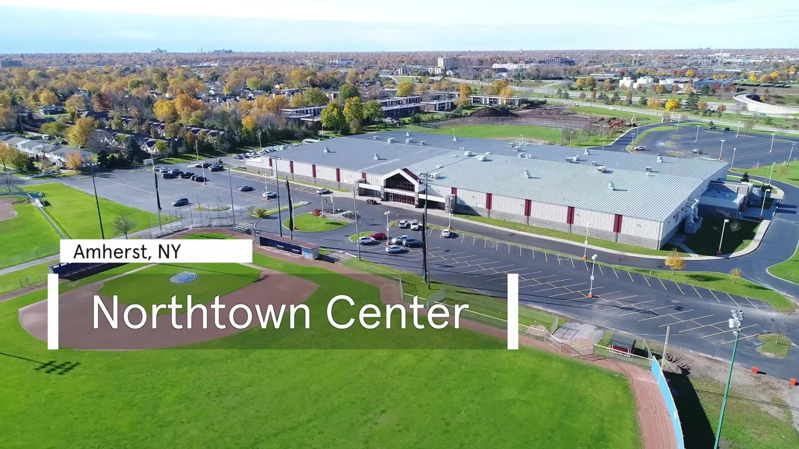 Aerial view of Northtown Center in autumn, its gabled glass entrance and red-striped metal siding beside the parking lot and a ball field. — Northtown Center at Amherst