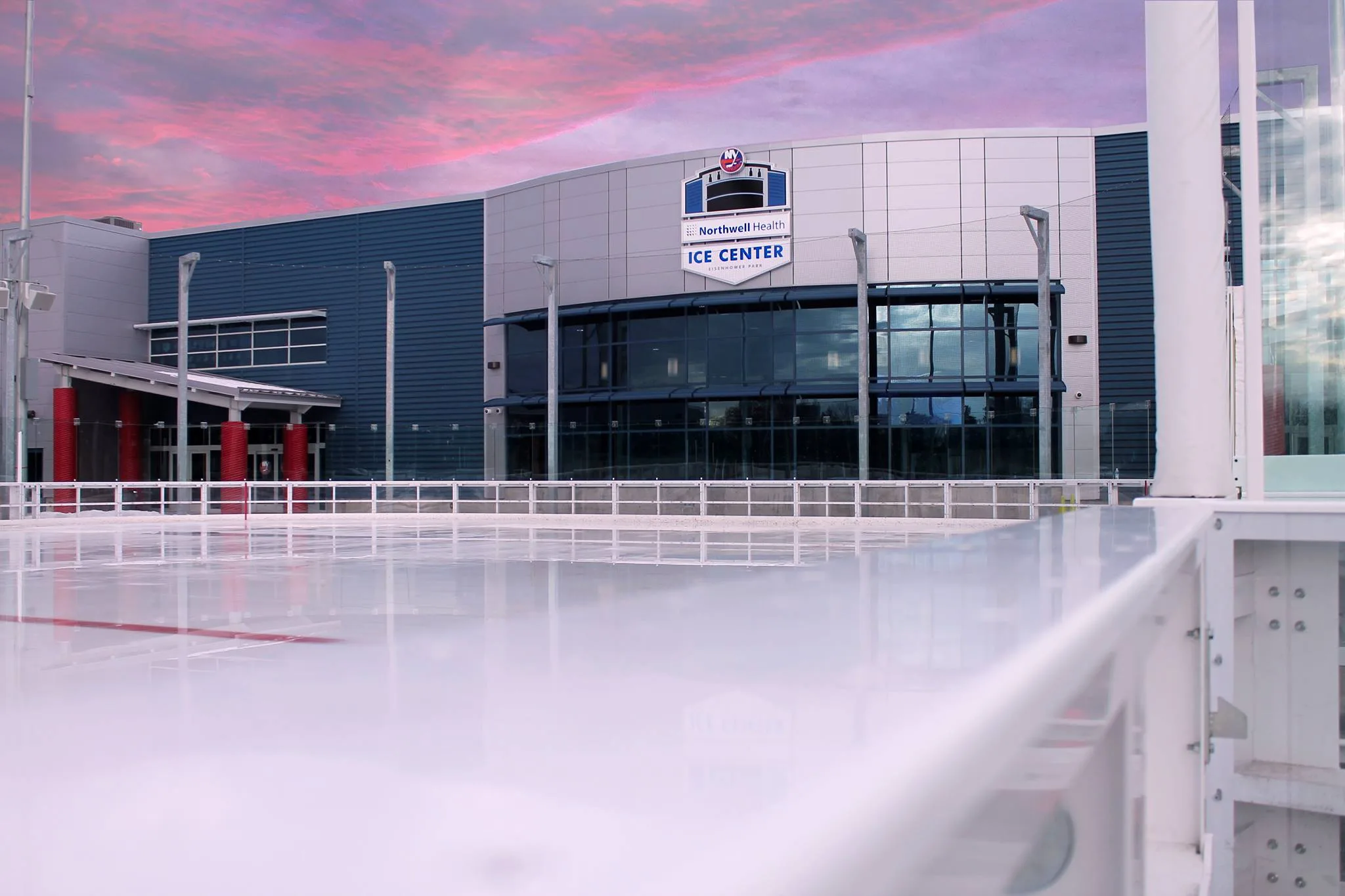Northwell Health Ice Center bajo un cielo rosado de atardecer, visto desde el otro lado de la pista exterior que tiene al frente — Northwell Health Ice Center