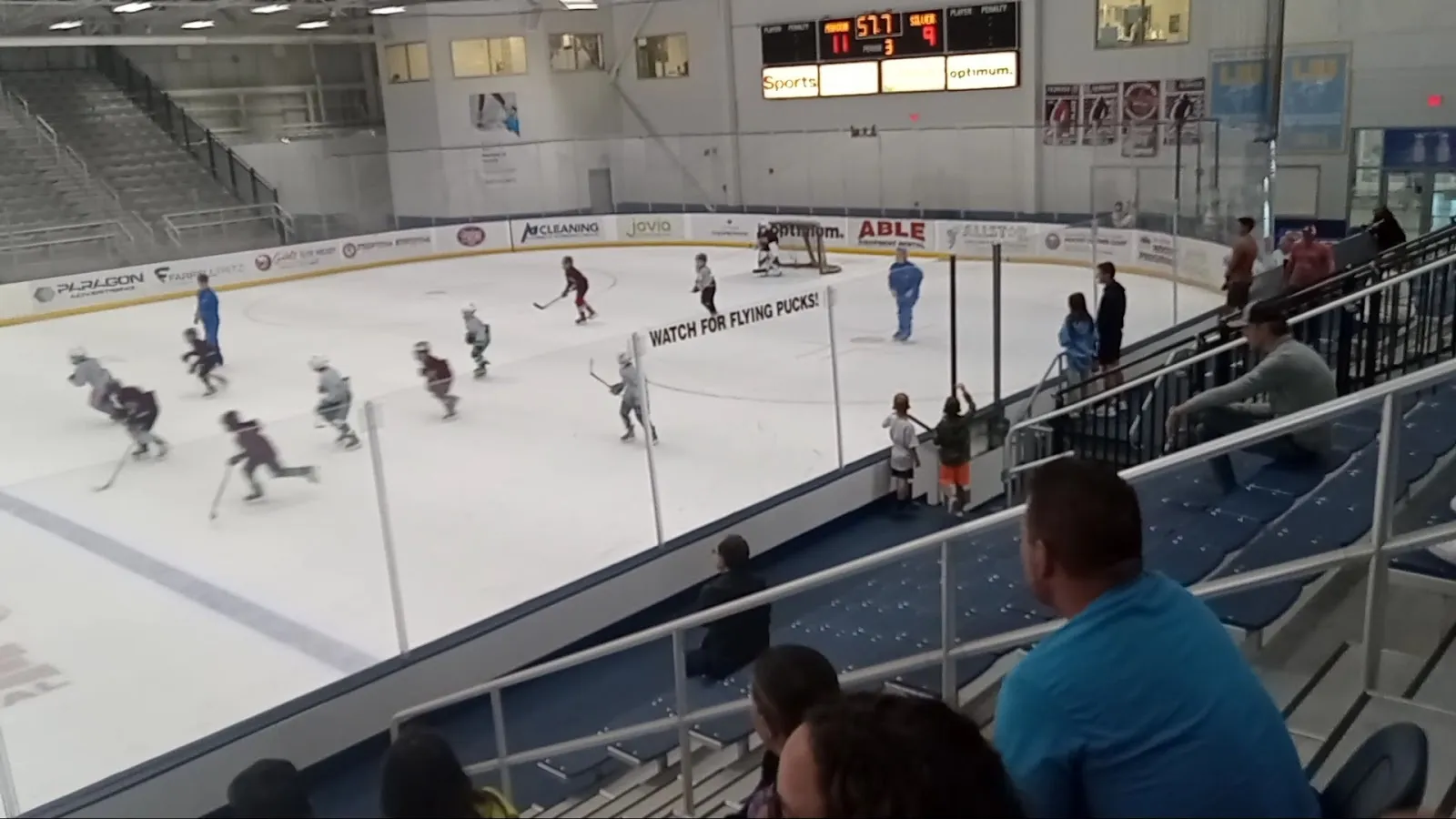 Un partido visto desde las gradas, con jugadores repartidos por el hielo blanco, el borde de unas gradas a la derecha y una pared de banderines detrás. — Northwell Health Ice Center