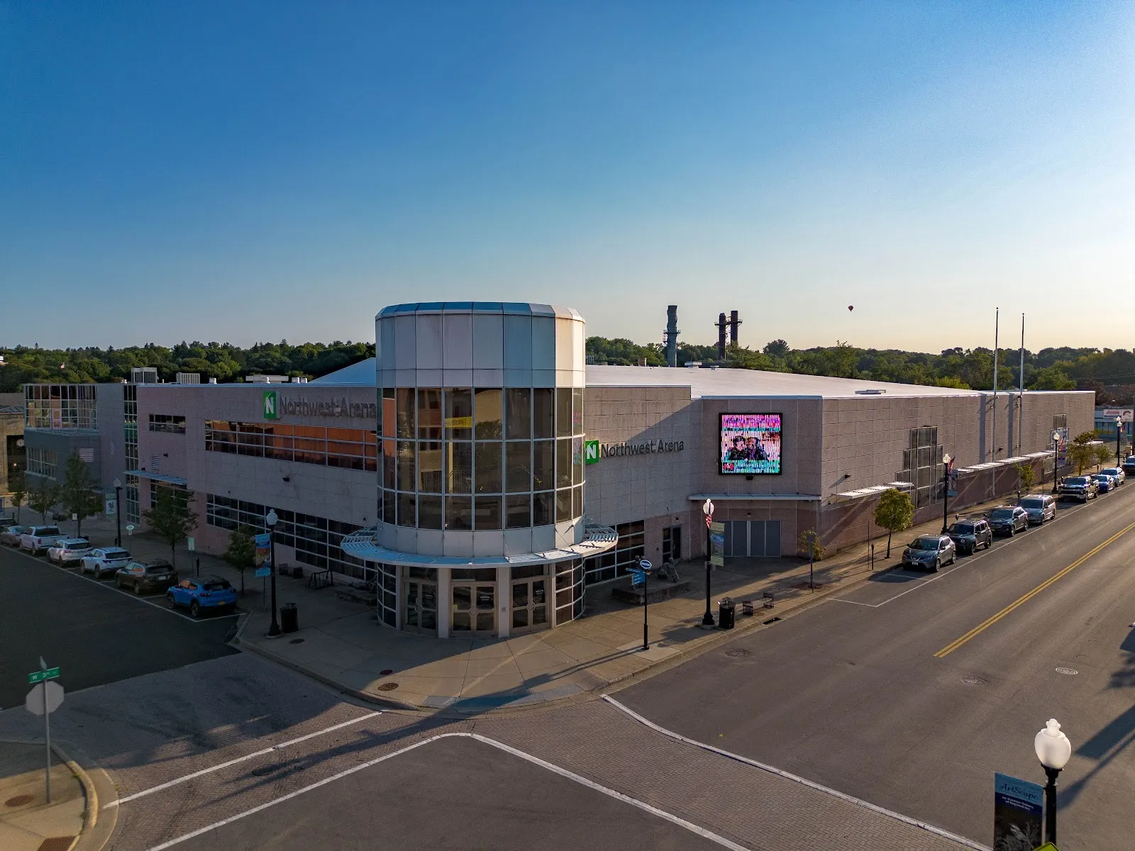 La torre esquinera de vidrio de la arena y el letrero 'Northwest Arena' en West Third Street, en el centro de Jamestown, con una pantalla de video en el muro lateral largo y autos estacionados junto a la banqueta. — Northwest Arena