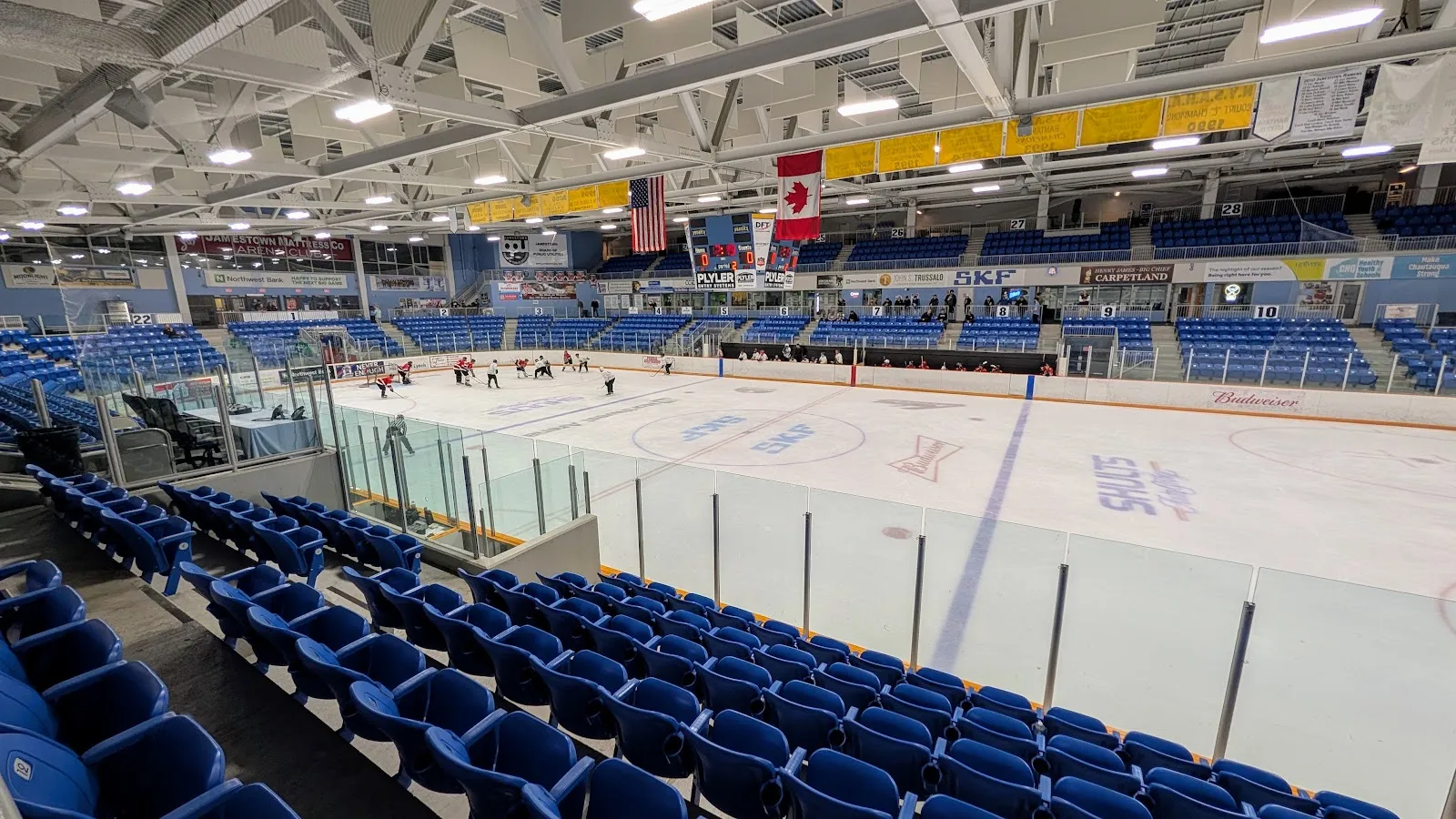 La pista principal desde los asientos azules, con mantas de campeonato y banderas de Estados Unidos y Canadá bajo las armaduras del techo, y un marcador central suspendido sobre el hielo. — Northwest Arena