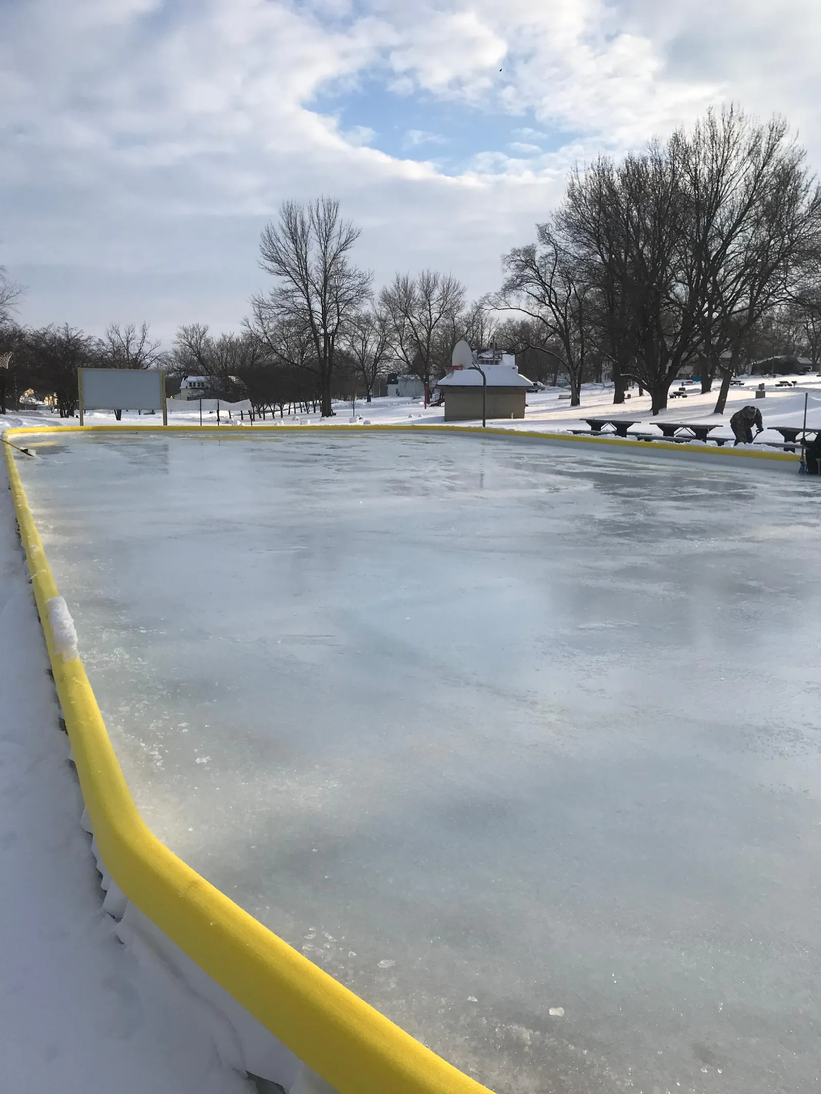 The Norwalk outdoor ice rink in winter, with its clear ice sheet in front and bare trees and a shelter beyond. — Norwalk Outdoor Ice Rink