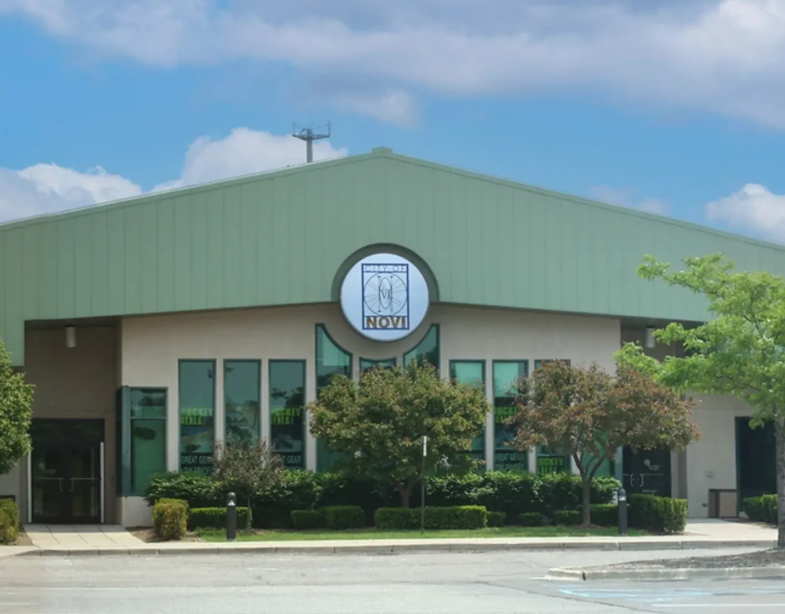 Front entrance of Novi Ice Arena, a green-roofed building with a circular NOVI logo sign above tall glass doors and windows. — Novi Ice Arena