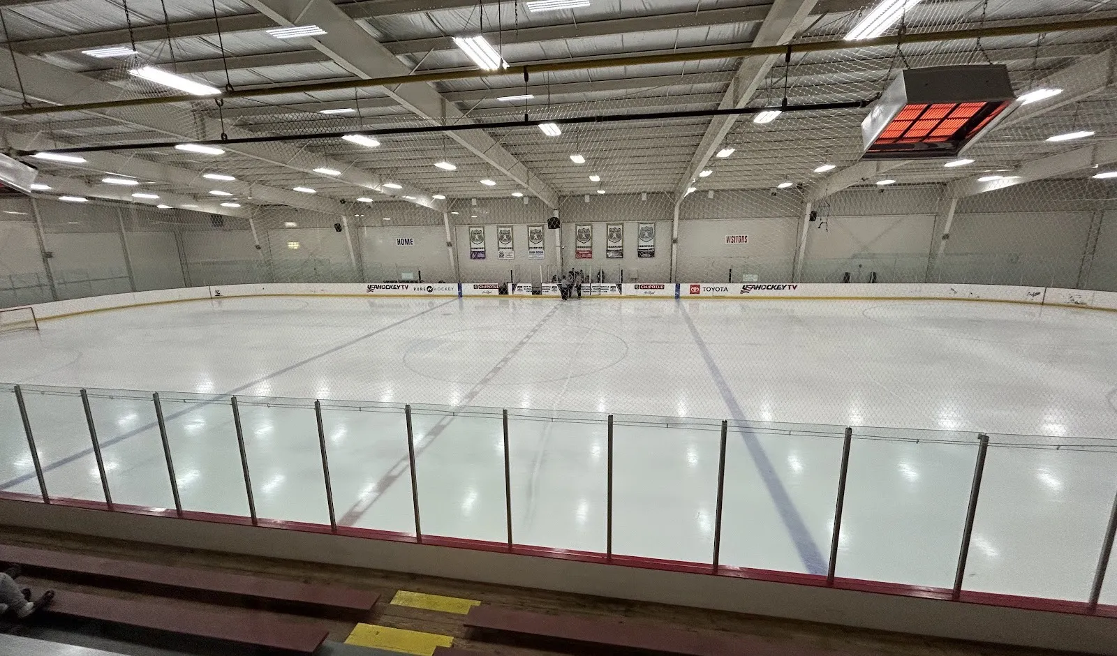 A Novi Ice Arena sheet from the bleachers, empty before a game, with the USA Hockey TV dasher boards and team banners on the far wall — Novi Ice Arena