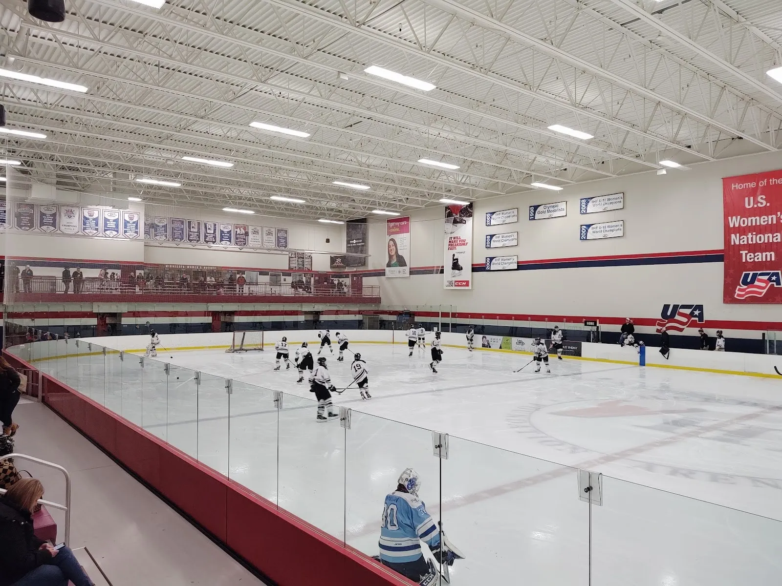 Una vista amplia a lo largo de la pista desde detrás de una esquina con barandal rojo, con jugadores repartidos por el hielo bajo un techo alto con banderines y anuncios de patrocinadores. — NSC Super Rink