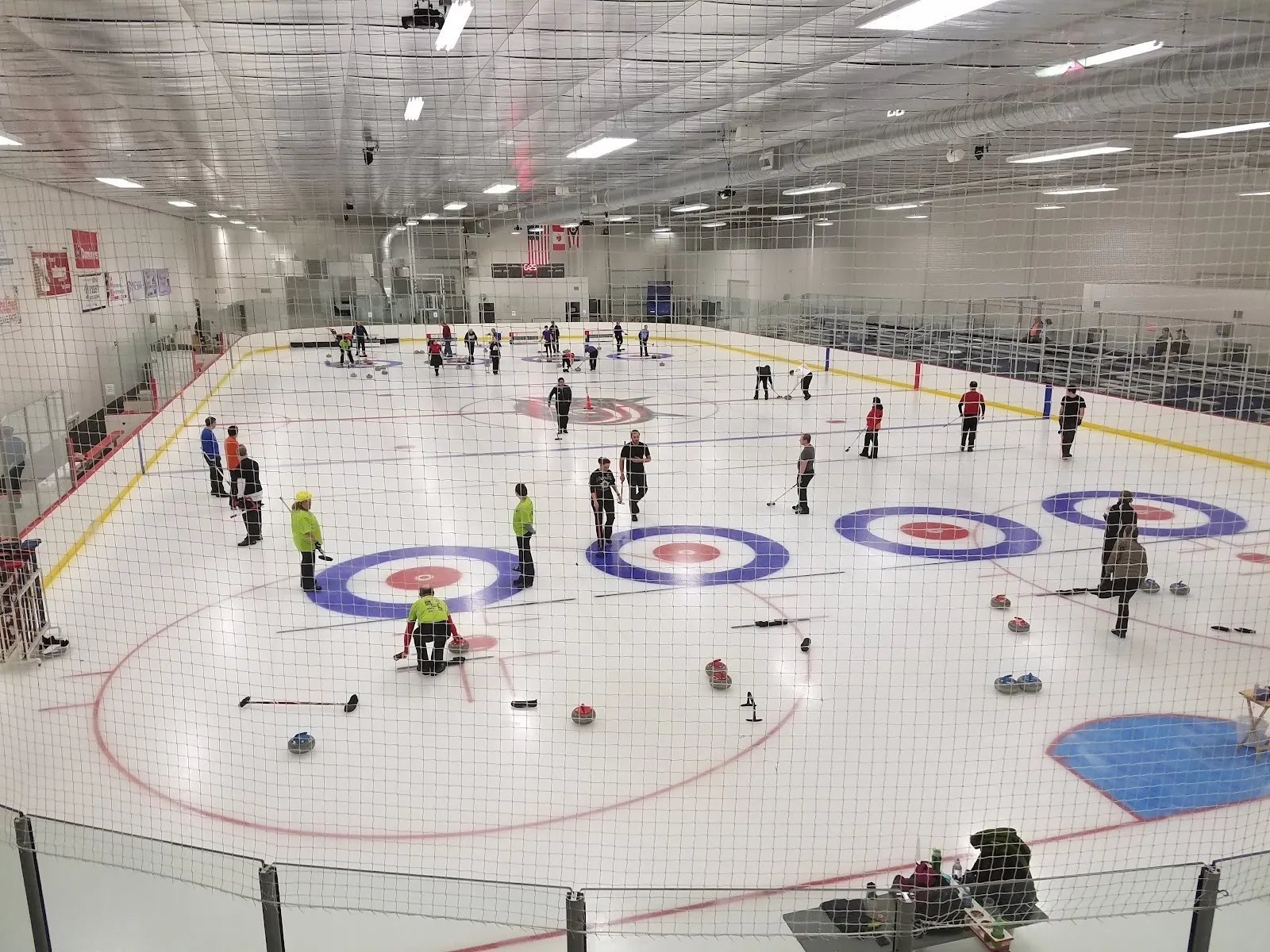 Una sesión de curling en una pista amplia, con personas repartidas por el hielo alrededor de varios círculos, con un techo de armaduras de acero y banderas sobre la pared del fondo. — NTPRD Chiller