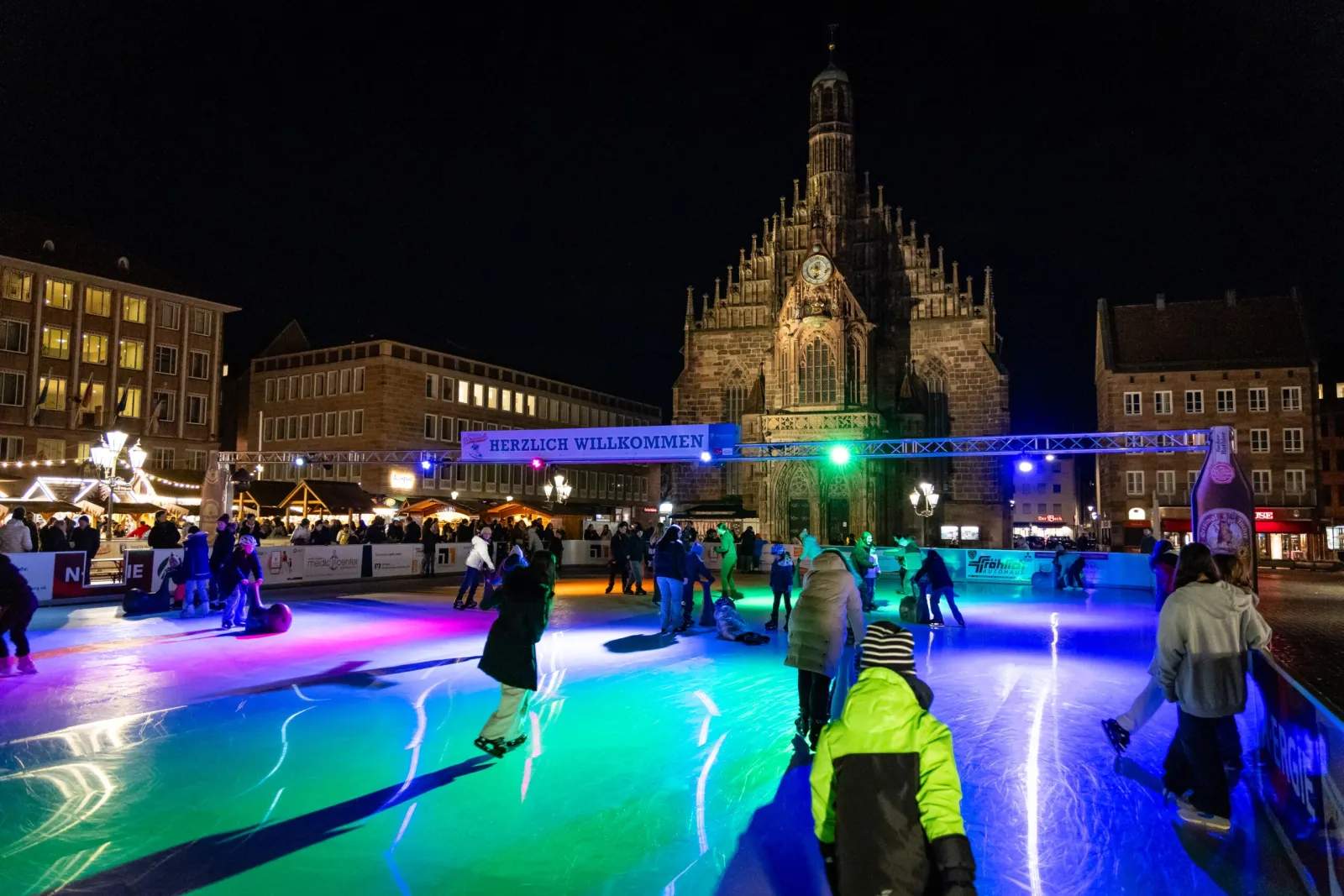 Die Eisbahn der Nürnberger Winterwelt auf dem Hauptmarkt bei Nacht, mit Läufern auf dem beleuchteten Eis und der Frauenkirche hinter dem Marktplatz. – Nürnberger Winterwelt