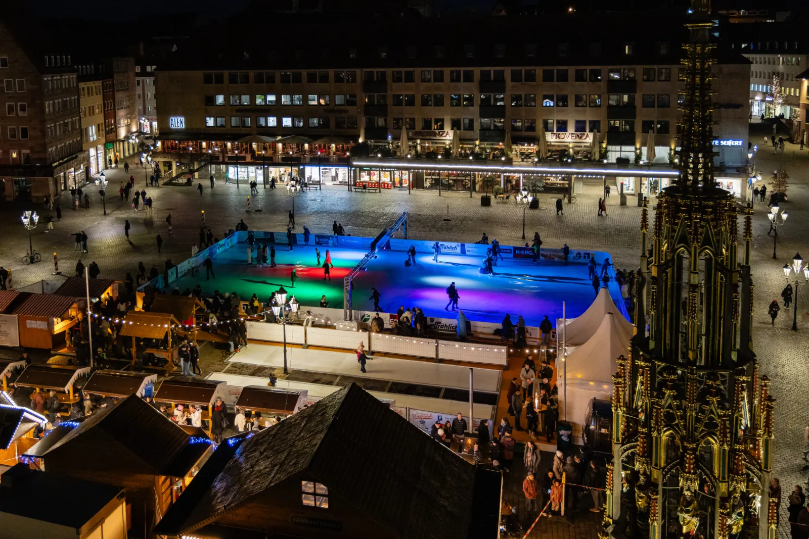 Die Nürnberger Winterwelt auf dem Hauptmarkt bei Nacht aus der Höhe, die beleuchtete Eisfläche mit Marktständen und einem Kirchturm daneben – Nürnberger Winterwelt