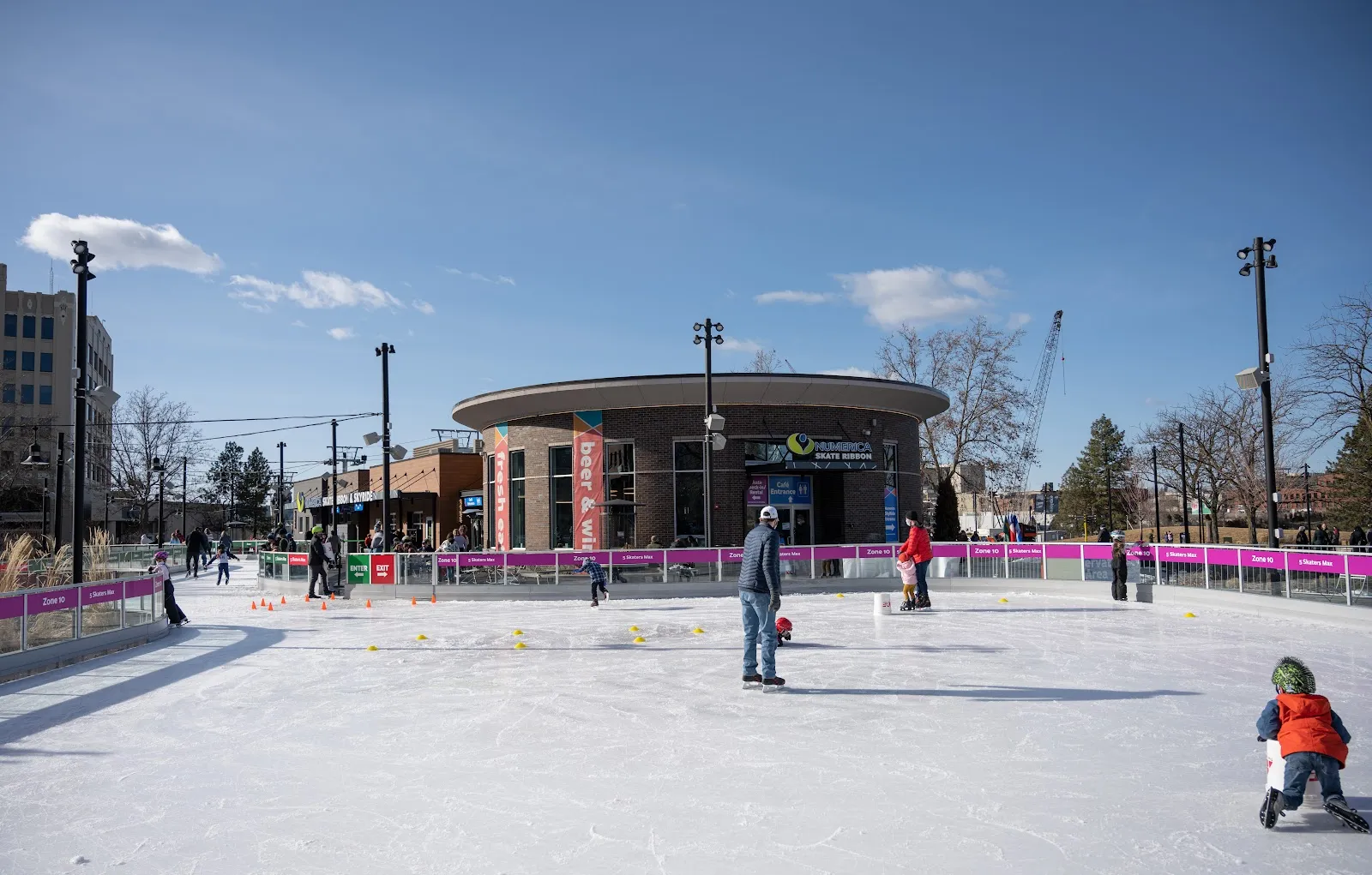 Skaters on the ribbon's ice in daylight, with the pavilion and downtown buildings behind — Numerica Skate Ribbon