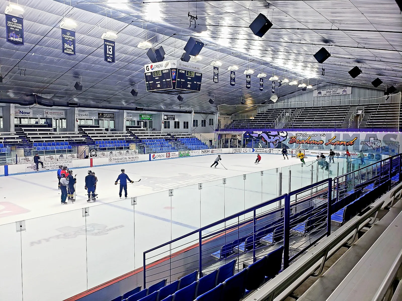 La pista vista desde las gradas detrás de las vallas, con unos cuantos patinadores cerca del extremo opuesto y las vigas y luces de la arena arriba. — NYTEX Sports Centre (North Richland Hills)
