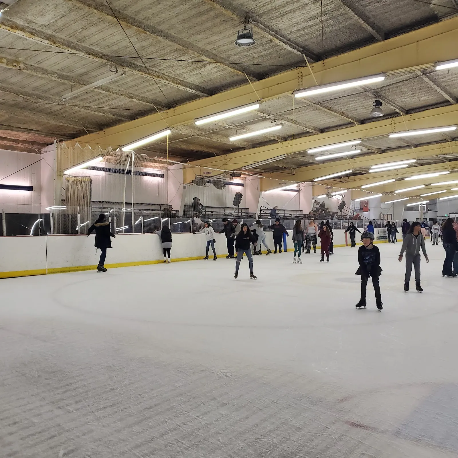 Wide interior view of the full ice sheet with a concrete beam ceiling, glass-and-boards rink surround, spectator bleachers, and a number of skaters on the ice during a public session. — Oak Park Ice Rink