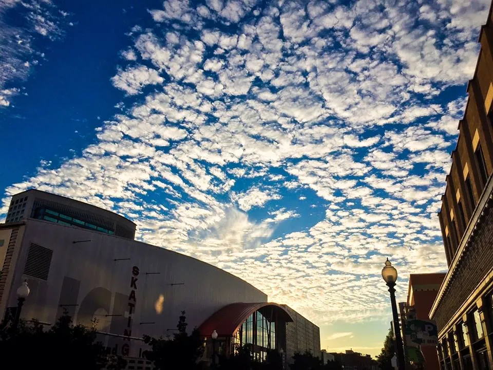 Dusk view looking up at the curved white rink building with a vertical 'SKATING' sign on its facade, street lamp and neighboring buildings alongside, dramatic cloud-streaked sky above. — Oakland Ice Center Operated by Sharks Ice