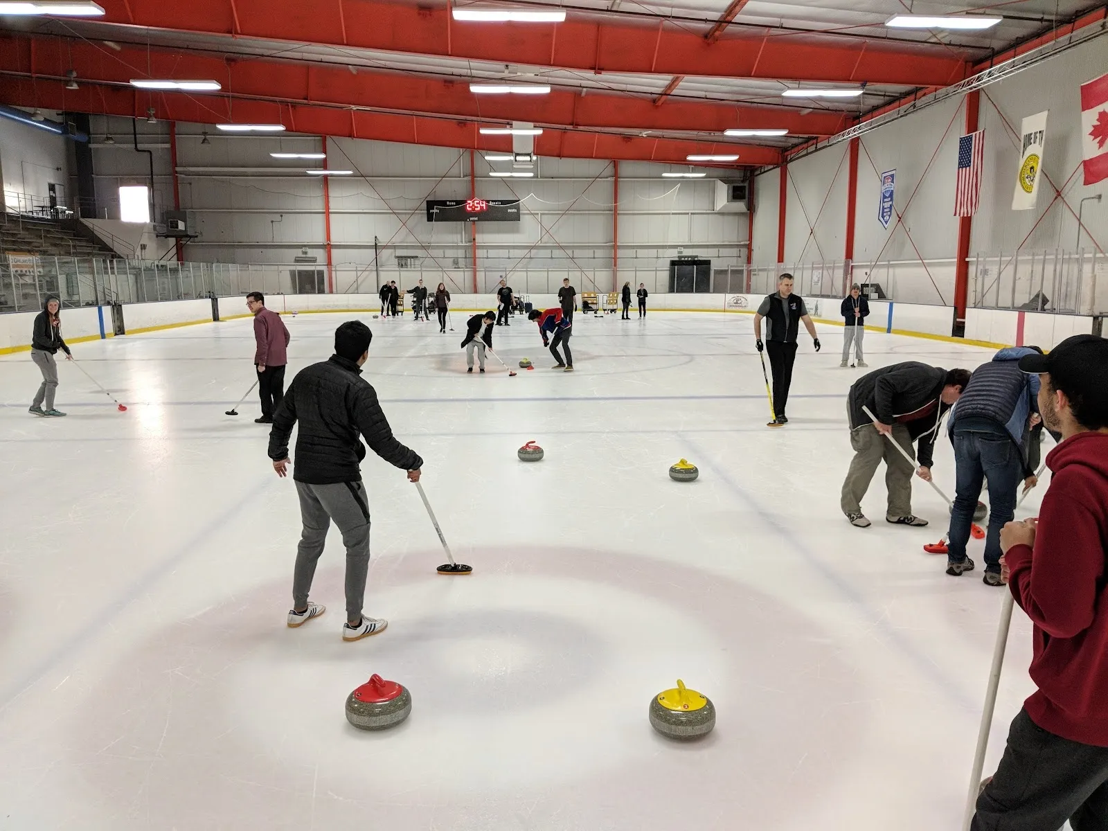 A curling session on the sheet, a skater bending over a stone among others spread across the ice under a red-trussed roof with banners on the far wall. — Oakland Ice Center Operated by Sharks Ice