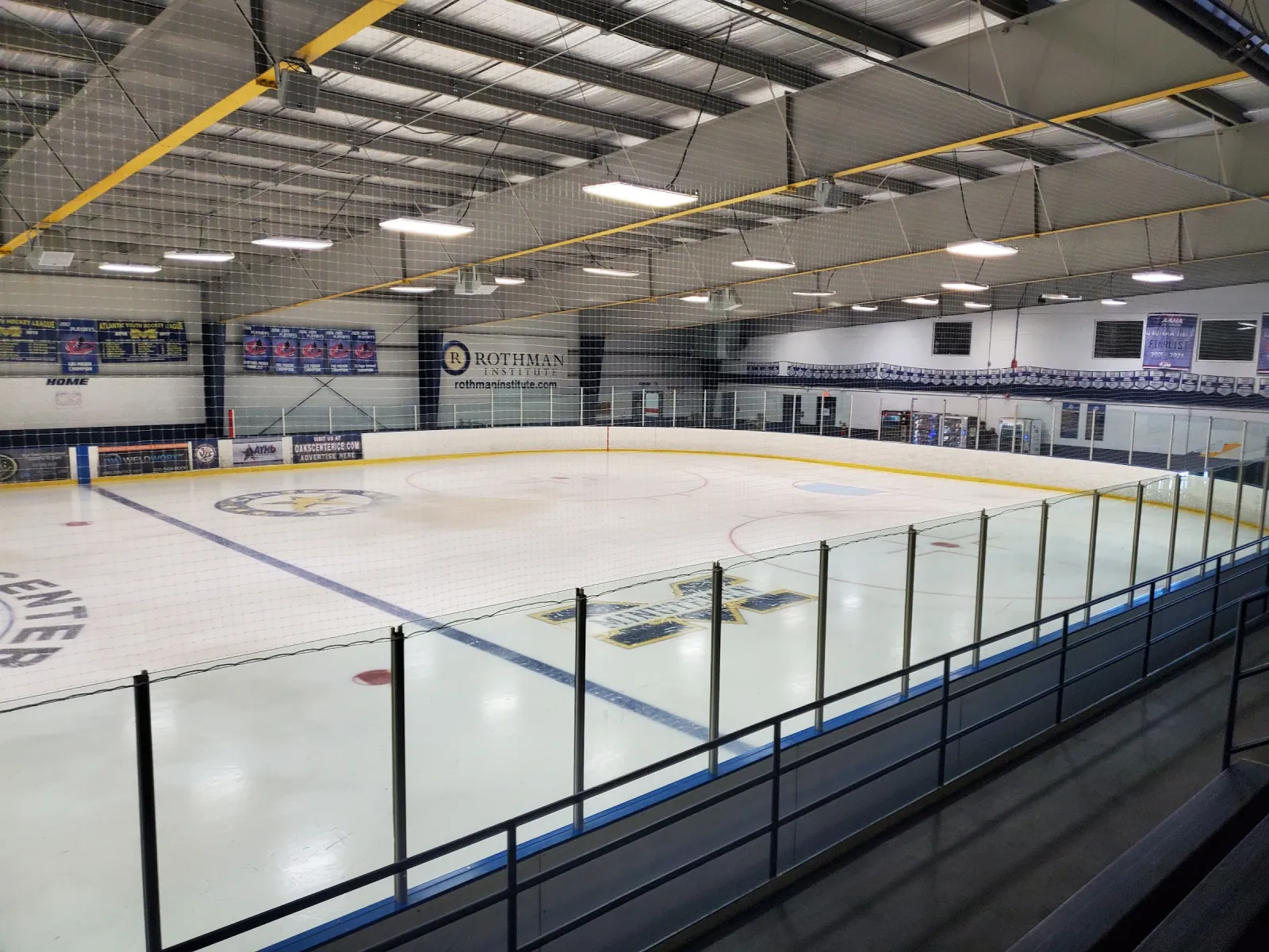 The ice seen from the stands behind the glass, with banners on the far wall and yellow roof beams overhead. — Oaks Center Ice