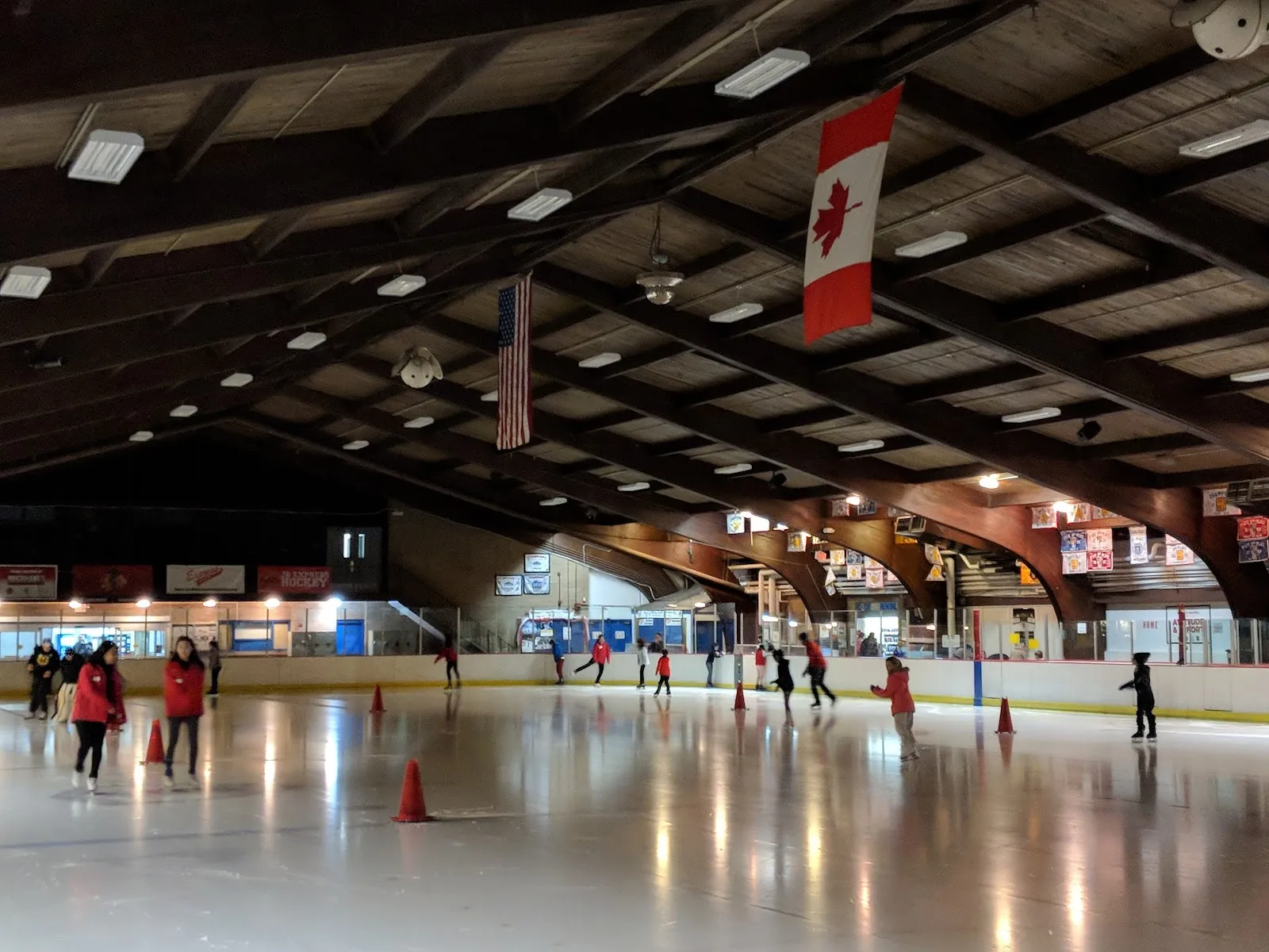 A public skating session under a dark timber roof, skaters spread across the ice among orange cones with Canadian and American flags on the end wall. — Oakton Sports Complex (Park Ridge)