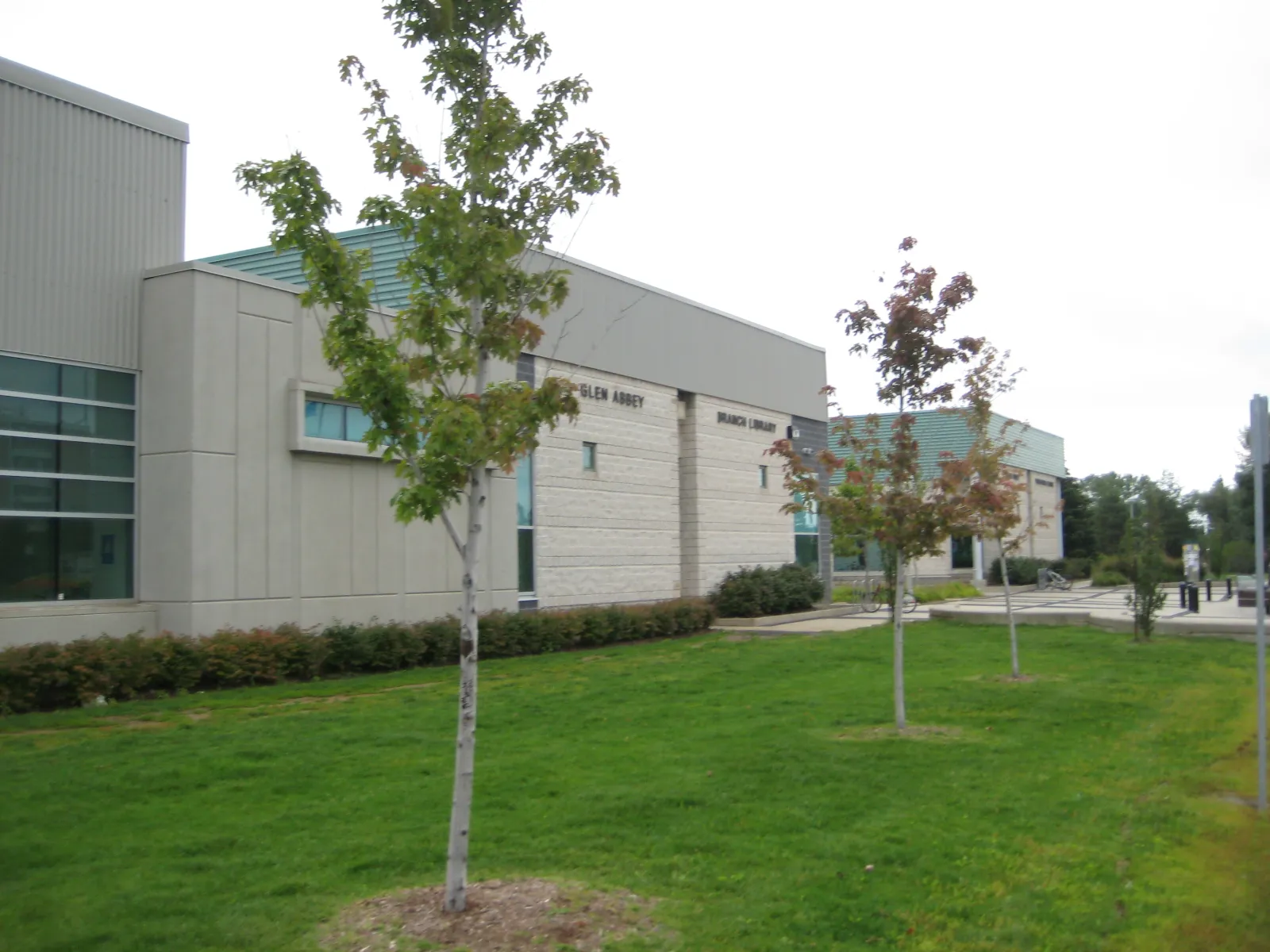 Exterior of the Glen Abbey Community Centre complex, showing the attached Glen Abbey Branch Library wing with beige brick cladding and a green standing-seam roof. — Glen Abbey Community Centre