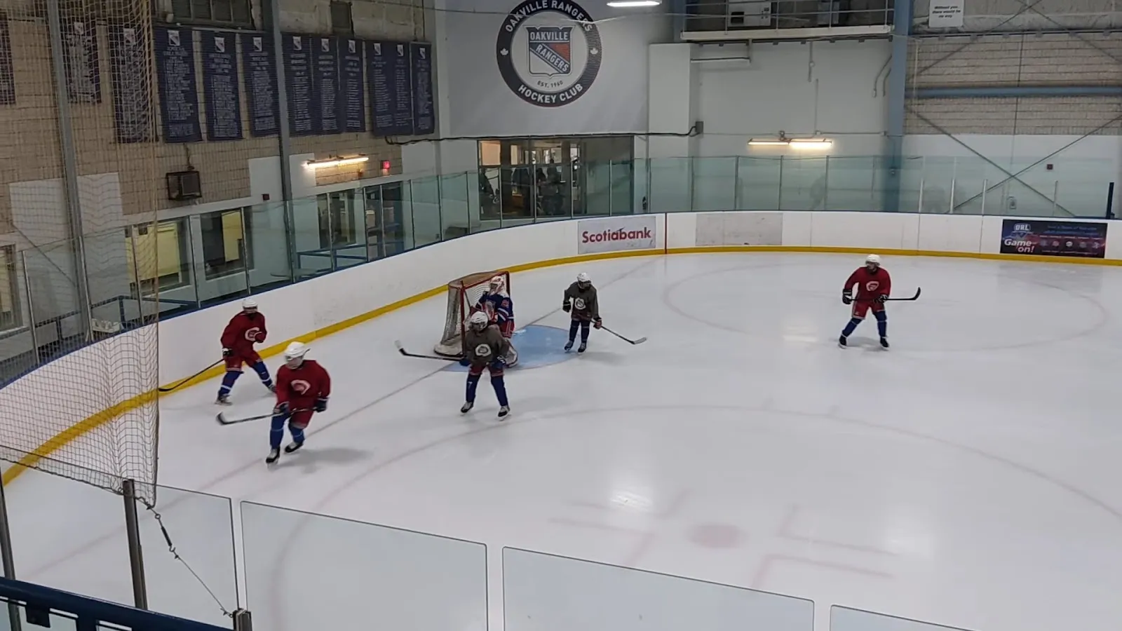 A game on a bright sheet seen through the glass rail, players spread along the far boards beneath a steel roof with a team banner at the left. — Glen Abbey Community Centre