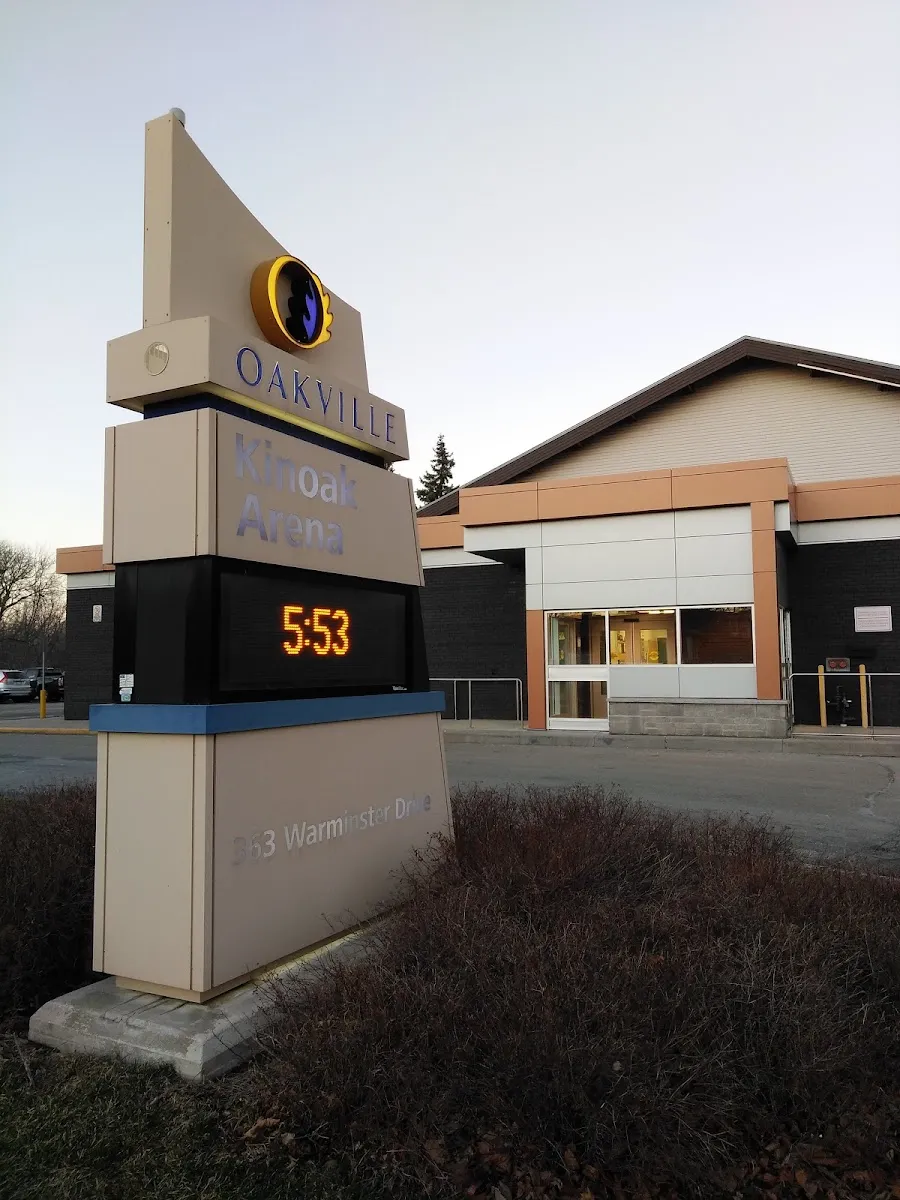 El letrero Oakville Kinoak Arena en 363 Warminster Drive, con un reloj electrónico, y detrás la entrada baja de ladrillo y paneles del edificio del arena al anochecer. — Kinoak Arena