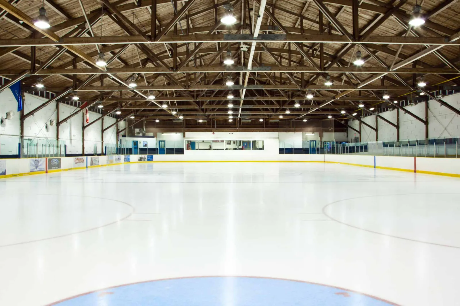 Pista de hielo vacía en Kinoak Arena, vista desde un extremo hacia las vallas, bajo un techo de armaduras de madera con la iluminación de la pista; no hay jugadores en el hielo. — Kinoak Arena