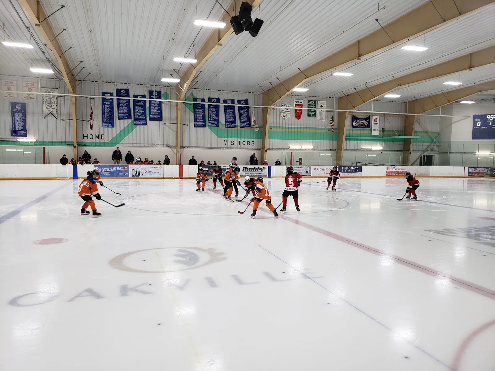 Un partido en el centro del hielo con un círculo con un escudo al estilo de los Oakville Rangers bajo un techo claro, con banderines de equipos en la pared del fondo. — River Oaks Community Centre