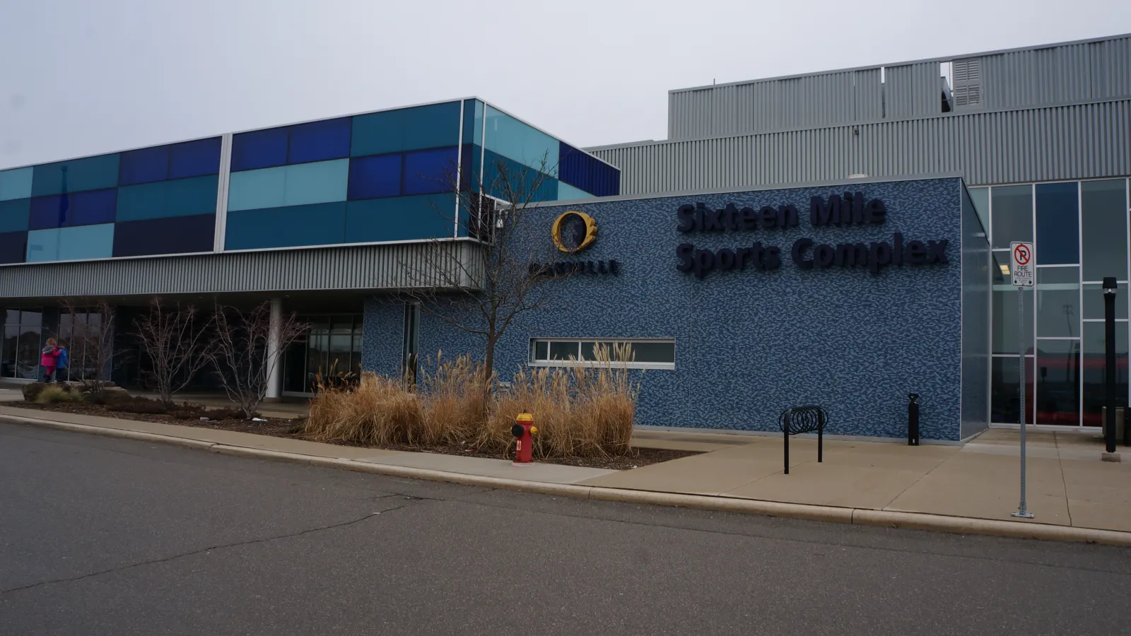 Entrada principal de Sixteen Mile Sports Complex, fachada de azulejo azul con el logotipo del Town of Oakville y letreros de "Sixteen Mile Sports Complex" en el edificio, un vestíbulo con muro cortina de vidrio y un hidrante al frente. — Sixteen Mile Sports Complex