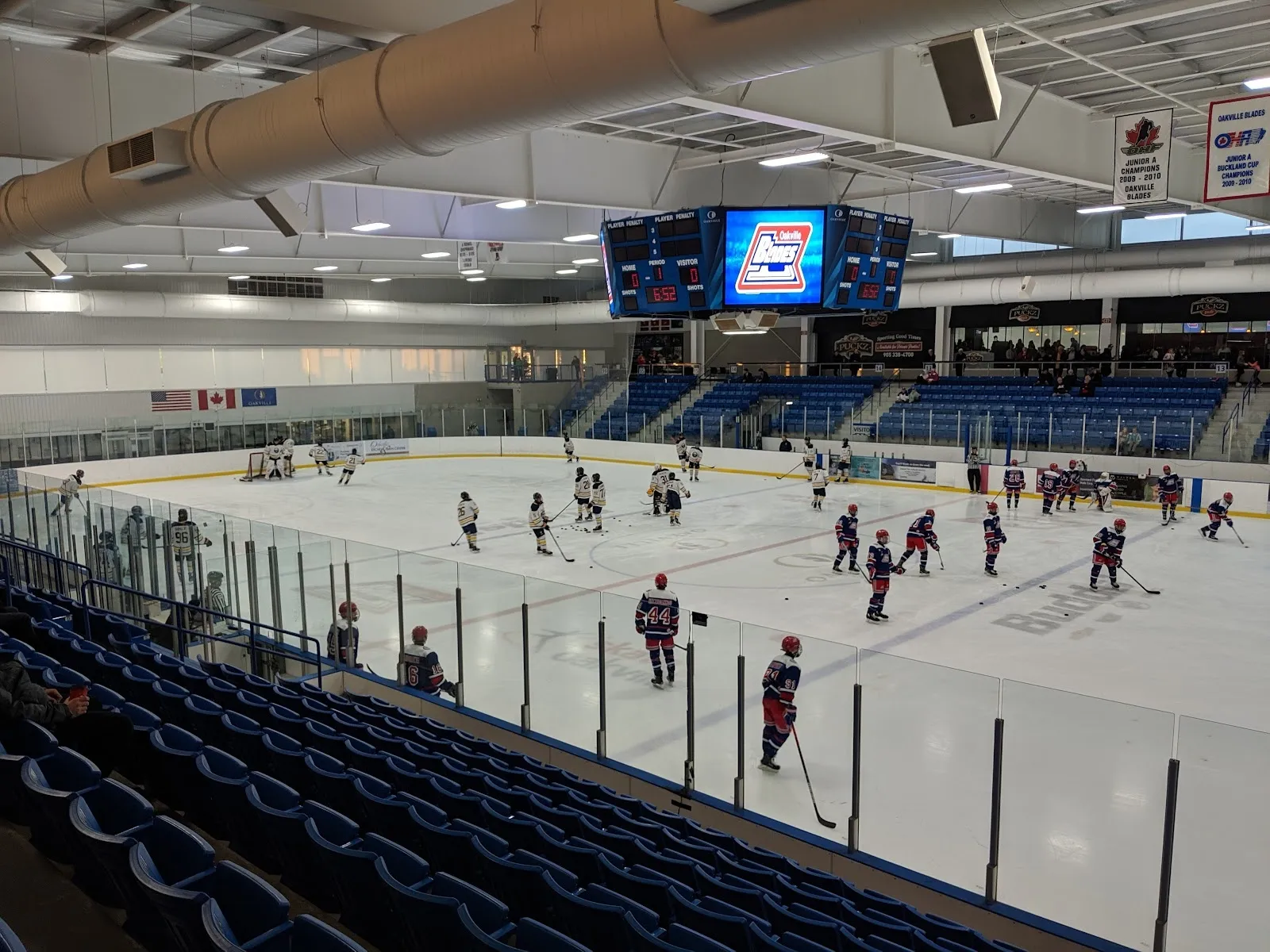 La arena desde las butacas bajas, con un partido a lo ancho de la pista, asientos azules alrededor y un marcador colgado sobre el centro del hielo. — Sixteen Mile Sports Complex