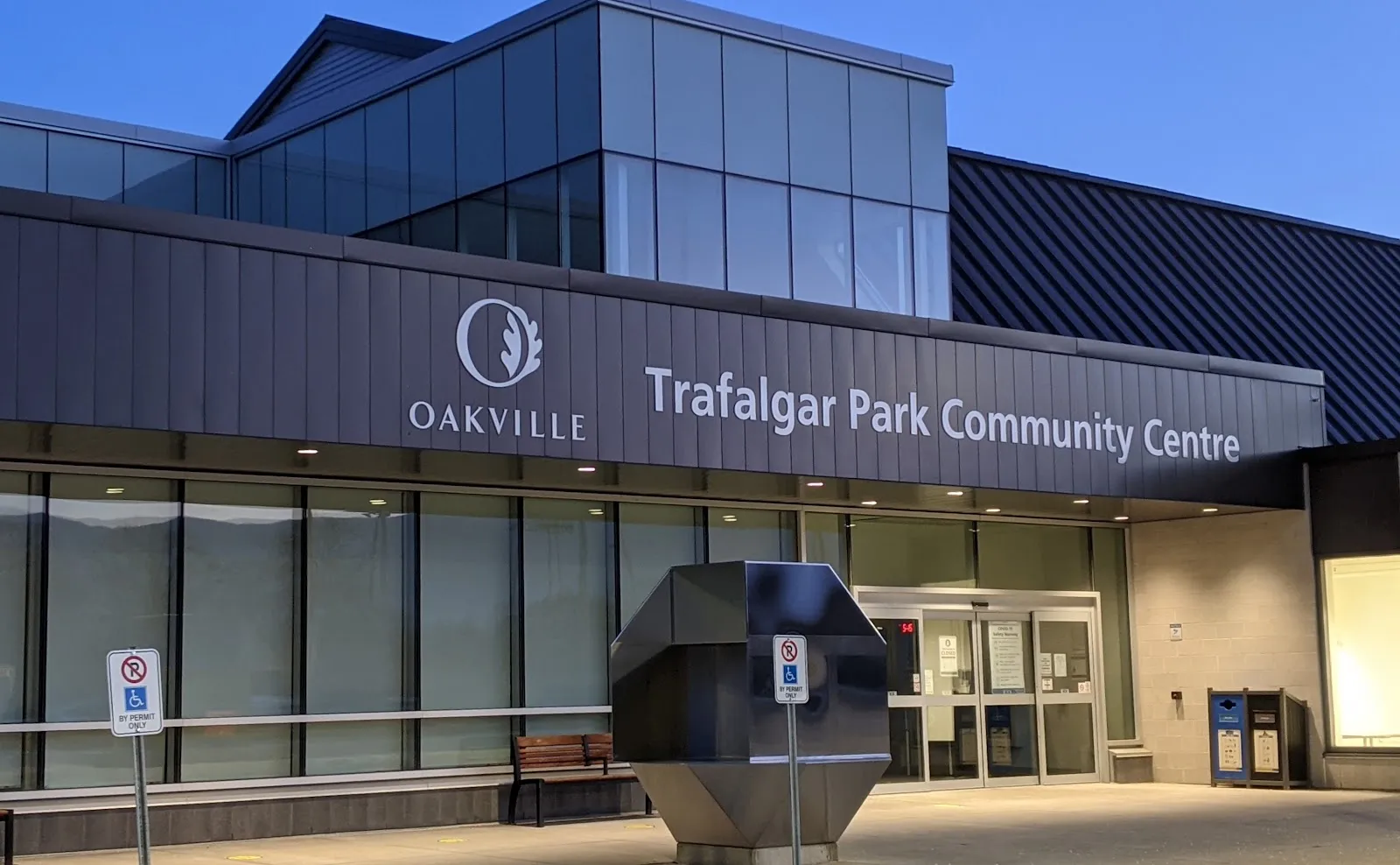 La entrada acristalada del Trafalgar Park Community Centre al anochecer, bajo el escudo de Oakville y el rótulo del centro, con las puertas correderas, un banco y señales de prohibido estacionar. — Trafalgar Park Community Centre