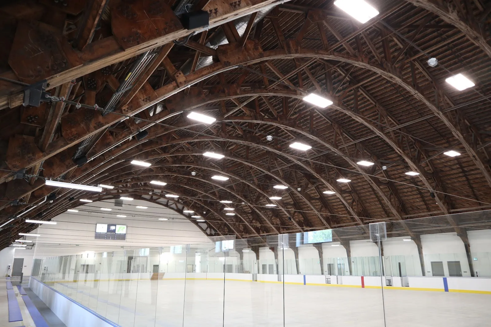 Toda la pista vacía del Trafalgar Park Community Centre bajo un histórico techo en arco de armaduras de madera, con vallas con cristal, líneas azules y amarillas y bancas a un lado. — Trafalgar Park Community Centre