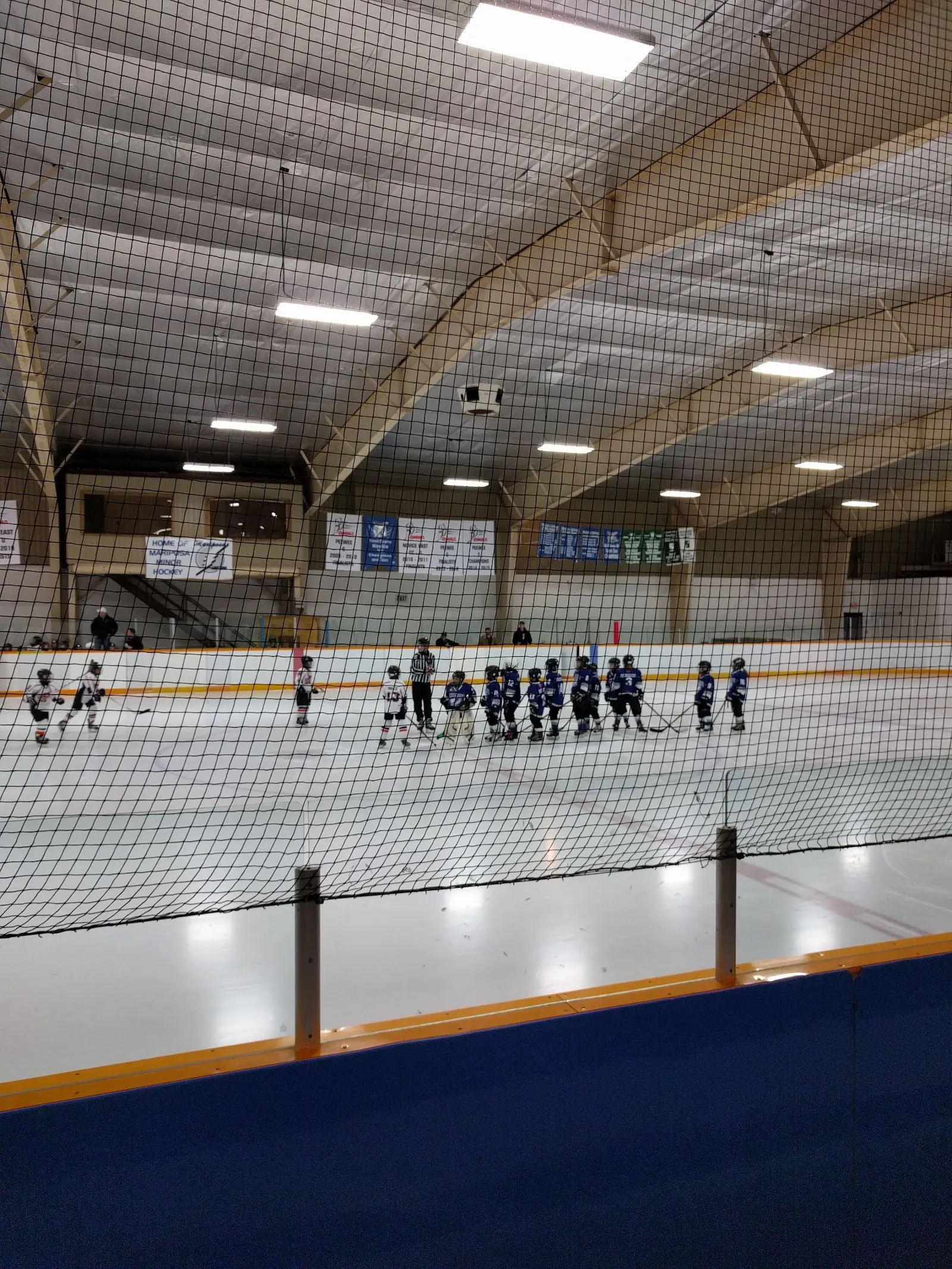 A row of young players lined up at the far side of the sheet behind netting, a timber-framed arched roof overhead with lights along the ridge. — Oakwood Community Centre (Kawartha Lakes)