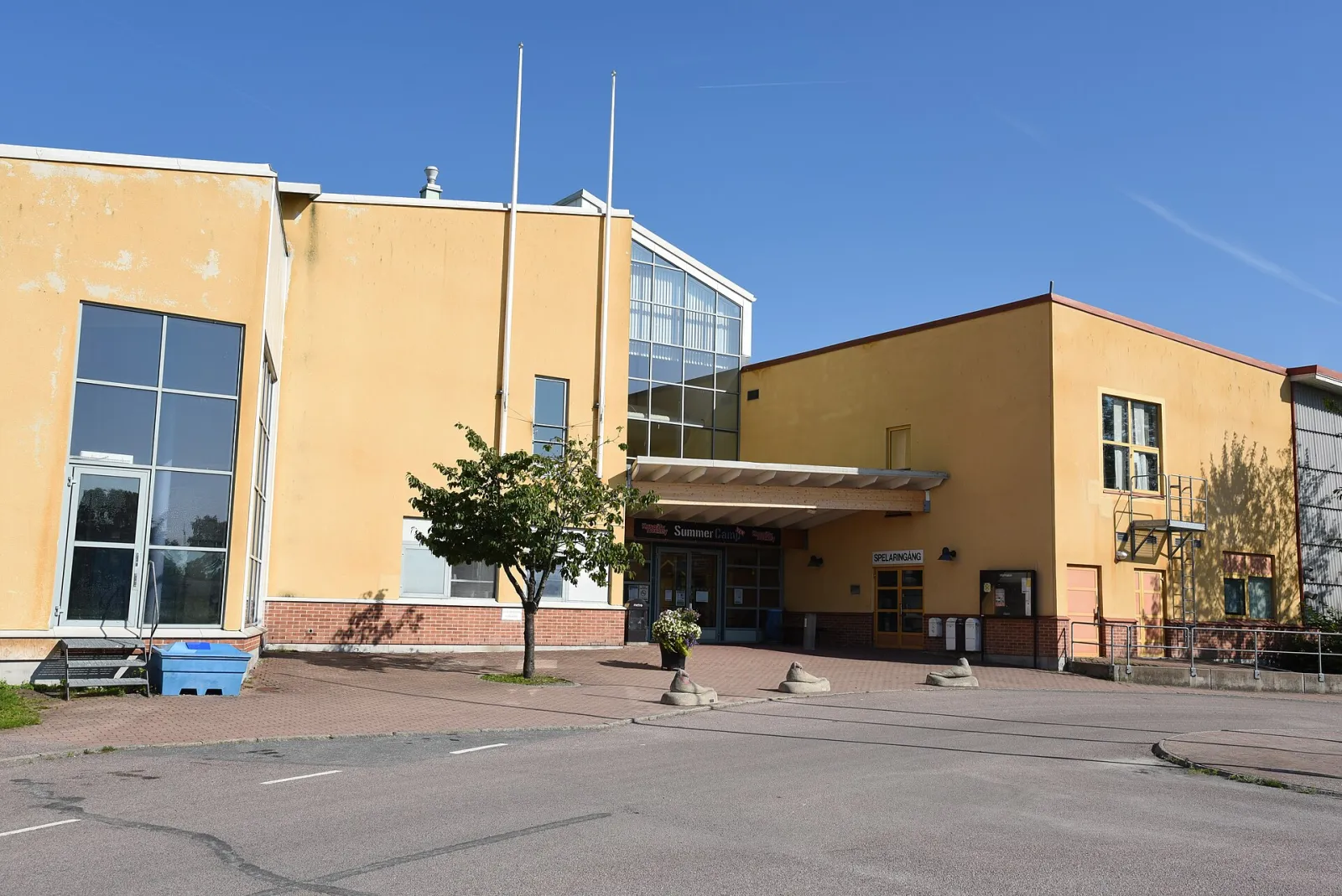 The yellow-rendered entrance front of Oasen sim- och ishall in Kungälv with its glazed gable, timber canopy over the doors, flagpoles and concrete seal sculptures in the forecourt. – Oasen sim- och ishall