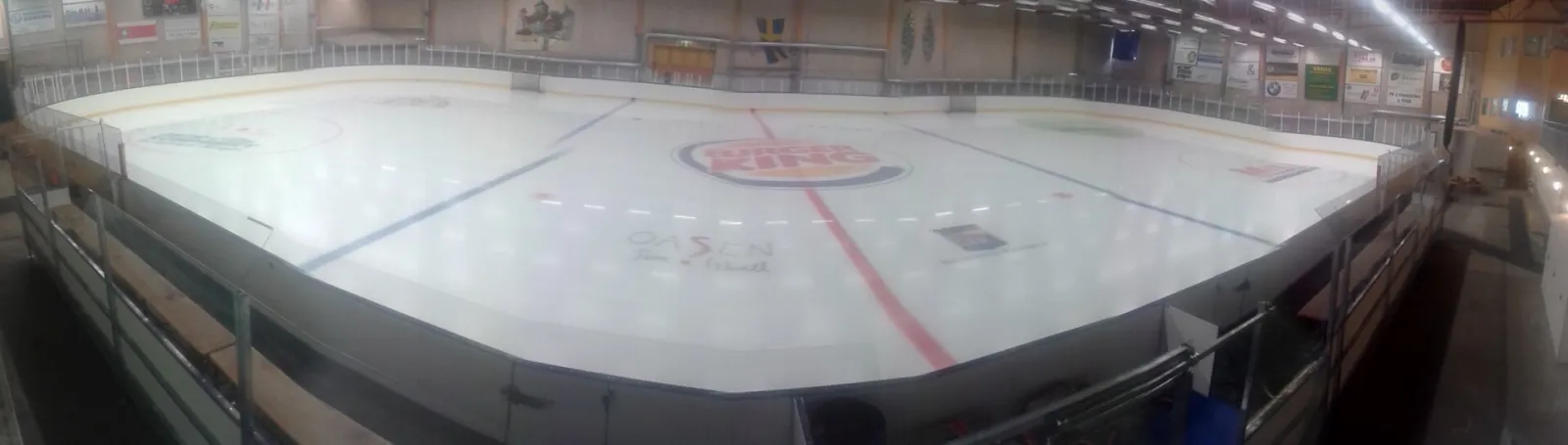 A wide-angle panorama of the empty ice sheet of the Kungälv ice hall at Oasen, with the centre-circle sponsor logo, red centre line, blue lines and the boards and bench side around it. – Oasen sim- och ishall