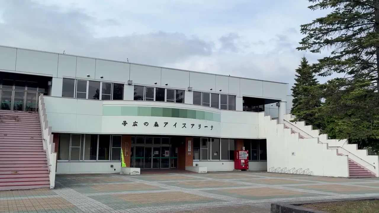 The front of Obihiro Forest Ice Arena with its green-banded two-storey entrance block and sign, stairs rising at both sides and a paved plaza in front — Obihiro Forest Ice Arena