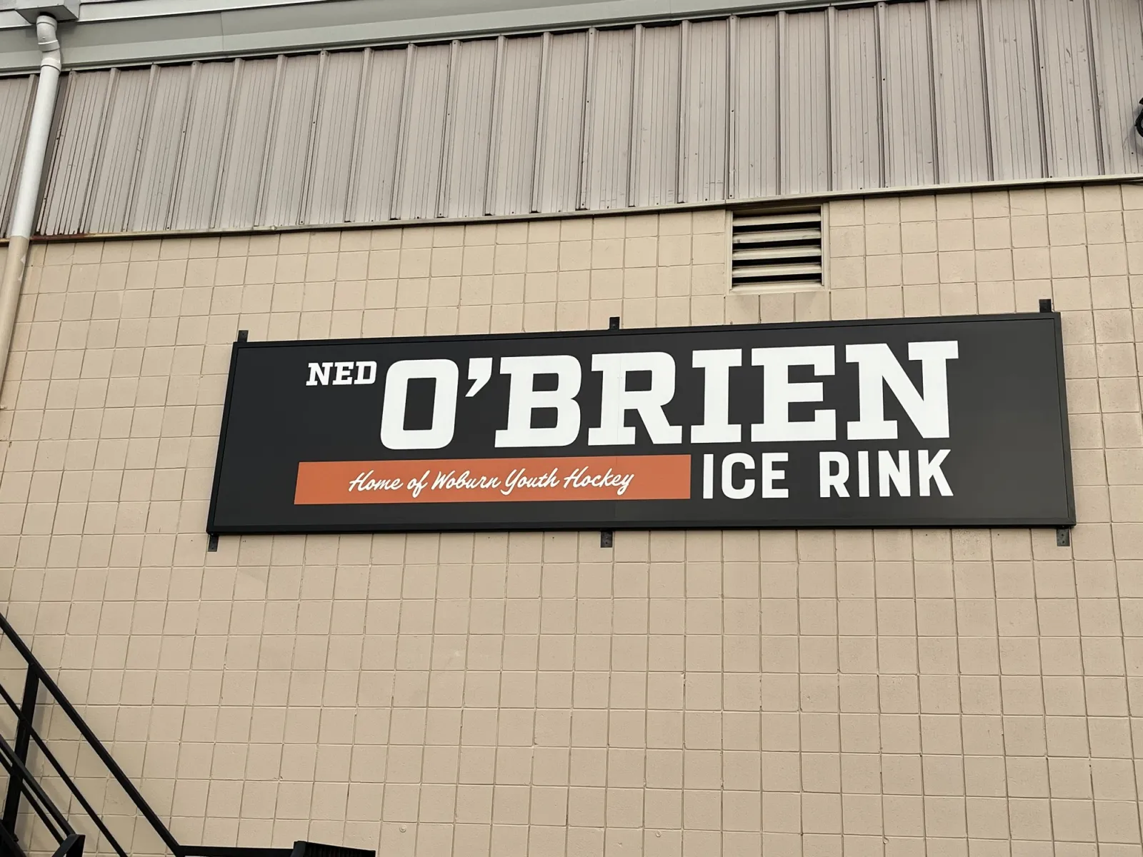 Building exterior with the black wall-mounted sign reading "NED O'BRIEN ICE RINK, Home of Woburn Youth Hockey" on the tan cinder-block facade below the rink's corrugated-metal roofline. — O'Brien Ice Rink