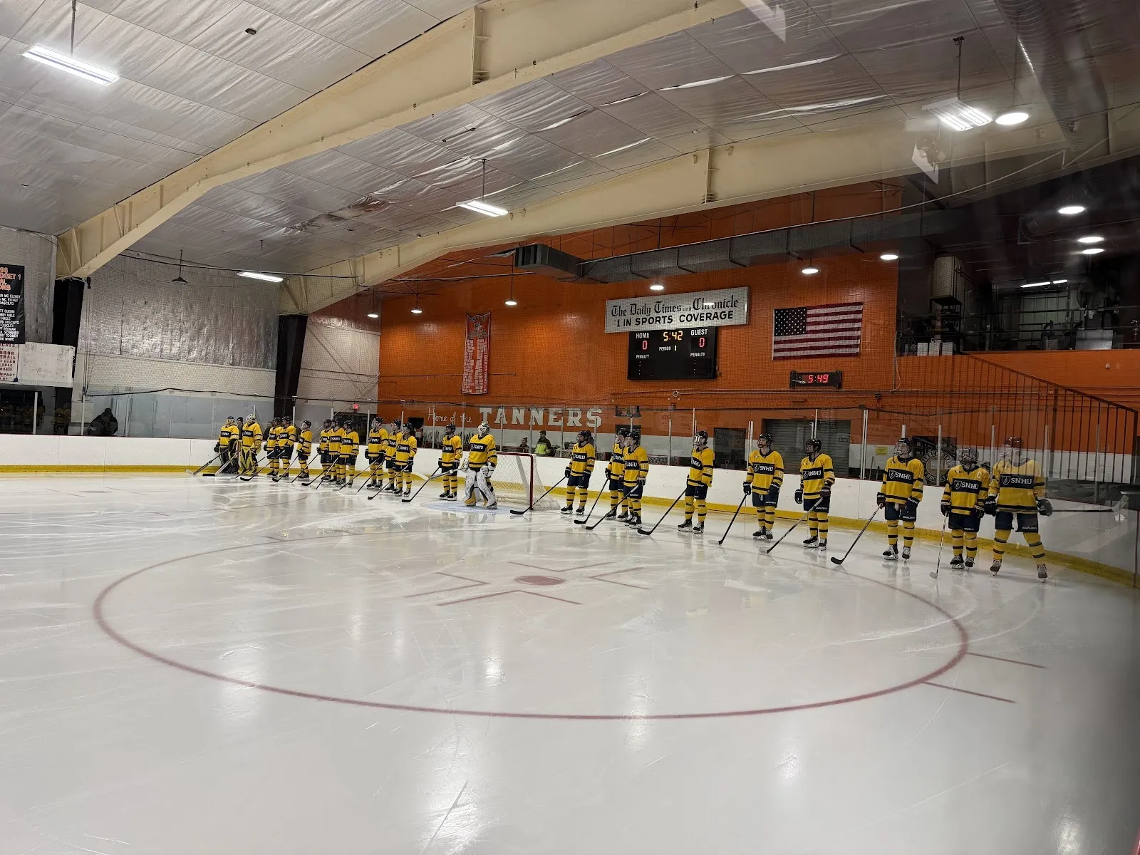 Two teams lined up on the blue lines of a wide sheet before a game, a red-circle faceoff dot at centre and an orange end wall with a flag behind. — O'Brien Ice Rink