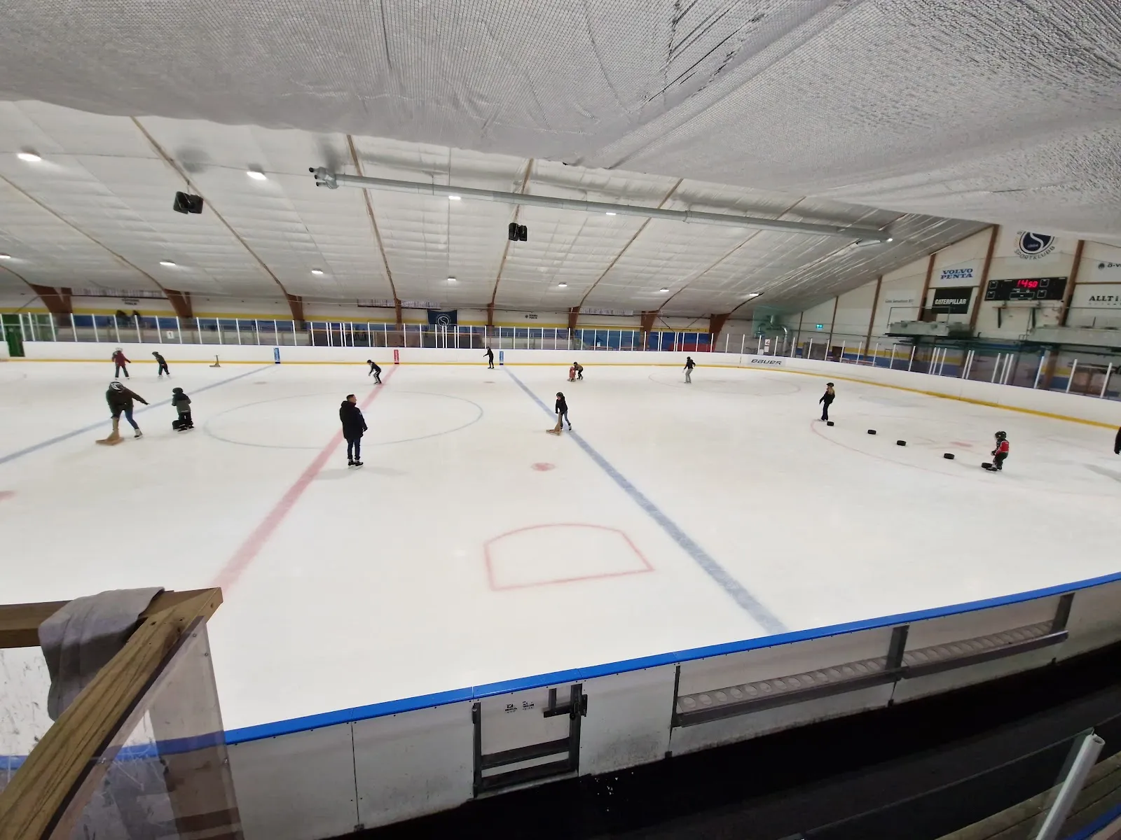 The ice sheet at Öckerö ishall seen from the stands, with blue-and-red lines, blue boards, the scoreboard and a curved timber roof, a few skaters small and spread across the ice — Öckerö ishall