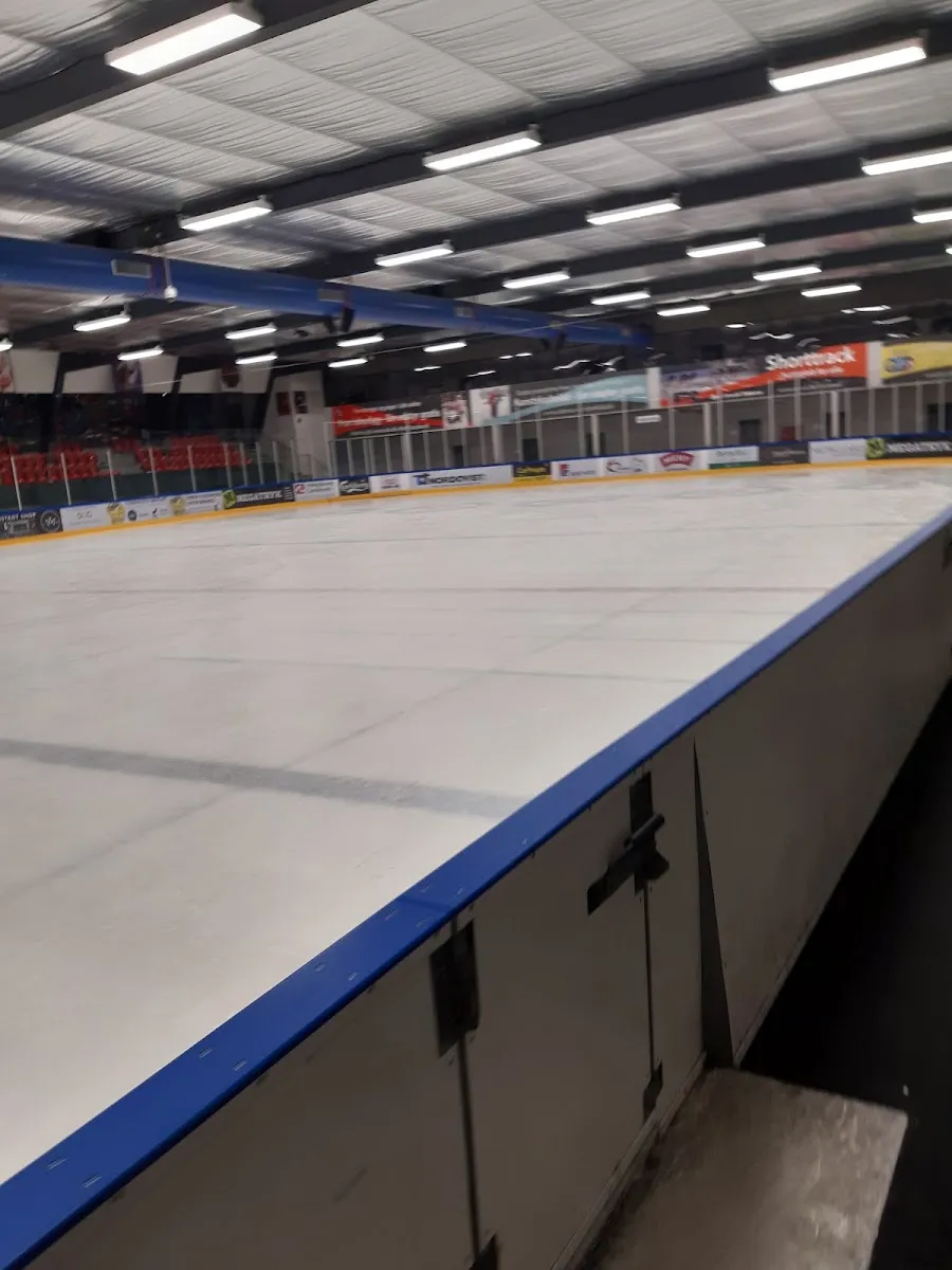 Den tomme isflade set langs banderne med blå kant, med det grå tag og lysstofrør over og sponsorpaneler og røde sæder for enden af hallen. — Odense Skøjtehal