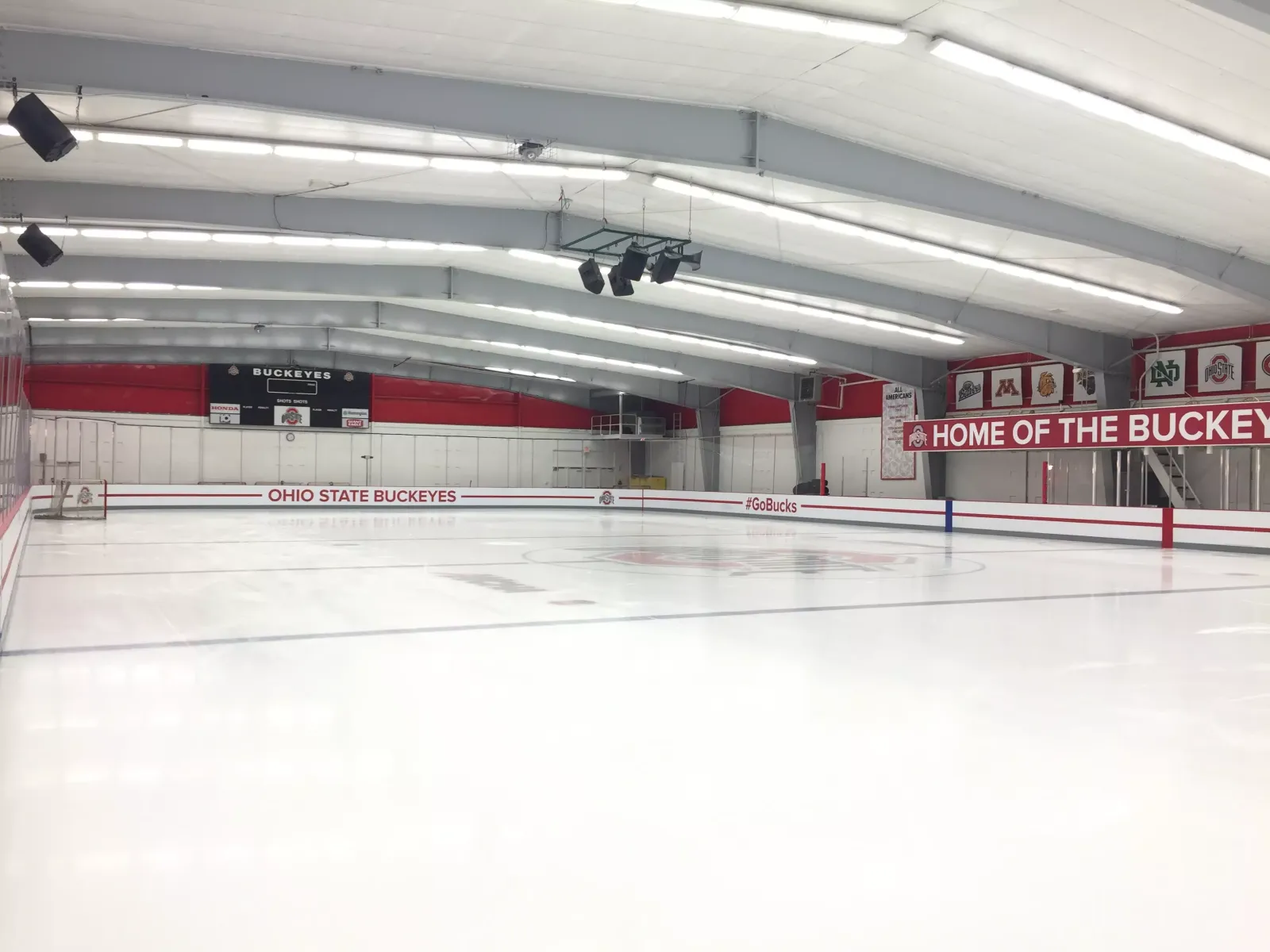 The empty sheet under rows of strip lights, with Ohio State Buckeyes lettering on the boards and a red Home of the Buckeyes banner on the far wall. — Ohio State Ice Rink