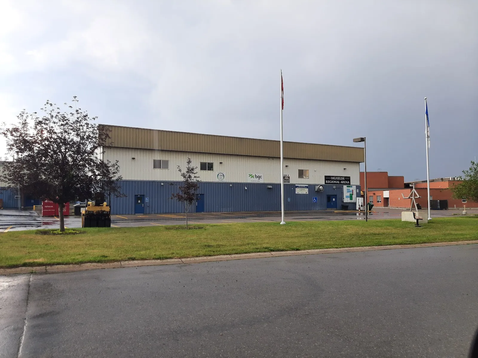 The blue-and-cream arena building behind a lawn with flagpoles, seen from the street on an overcast day. — Oilfields Regional Arena