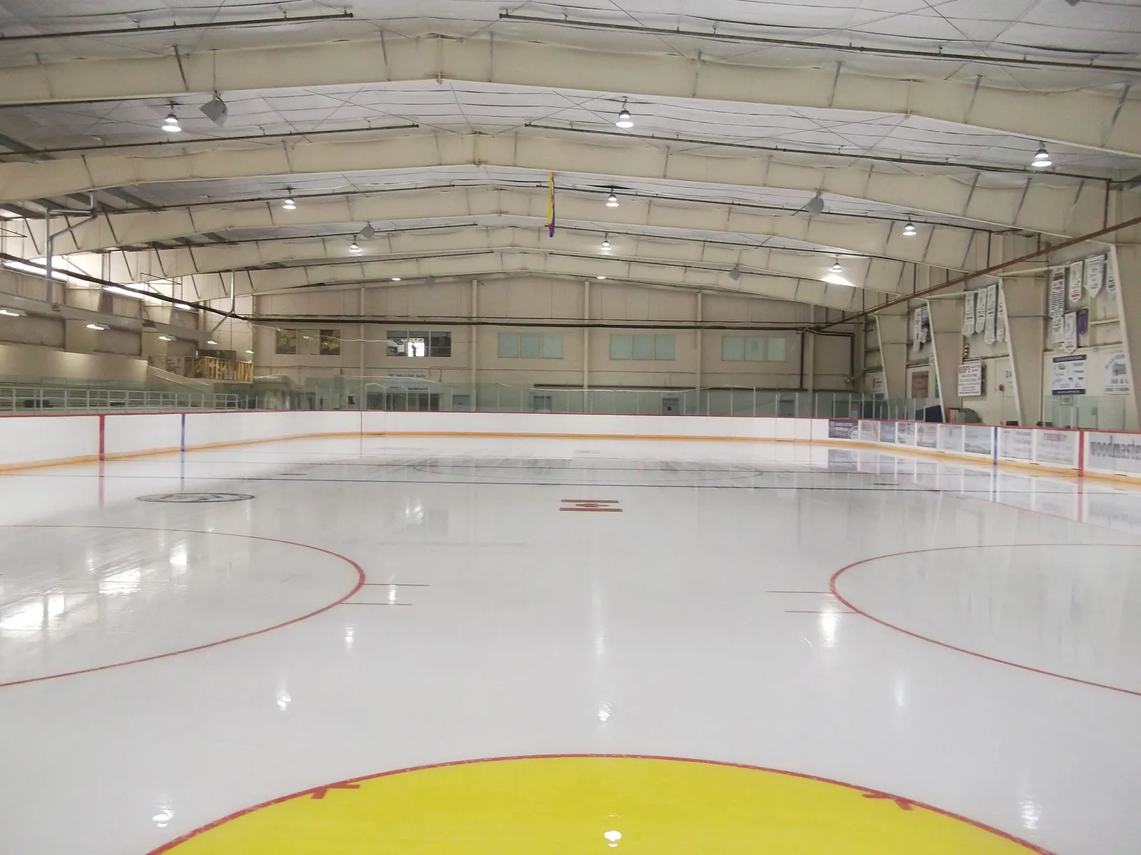 The empty indoor sheet under a white arched roof, with a yellow centre circle in the foreground and the stands along the left side. — Oilfields Regional Arena