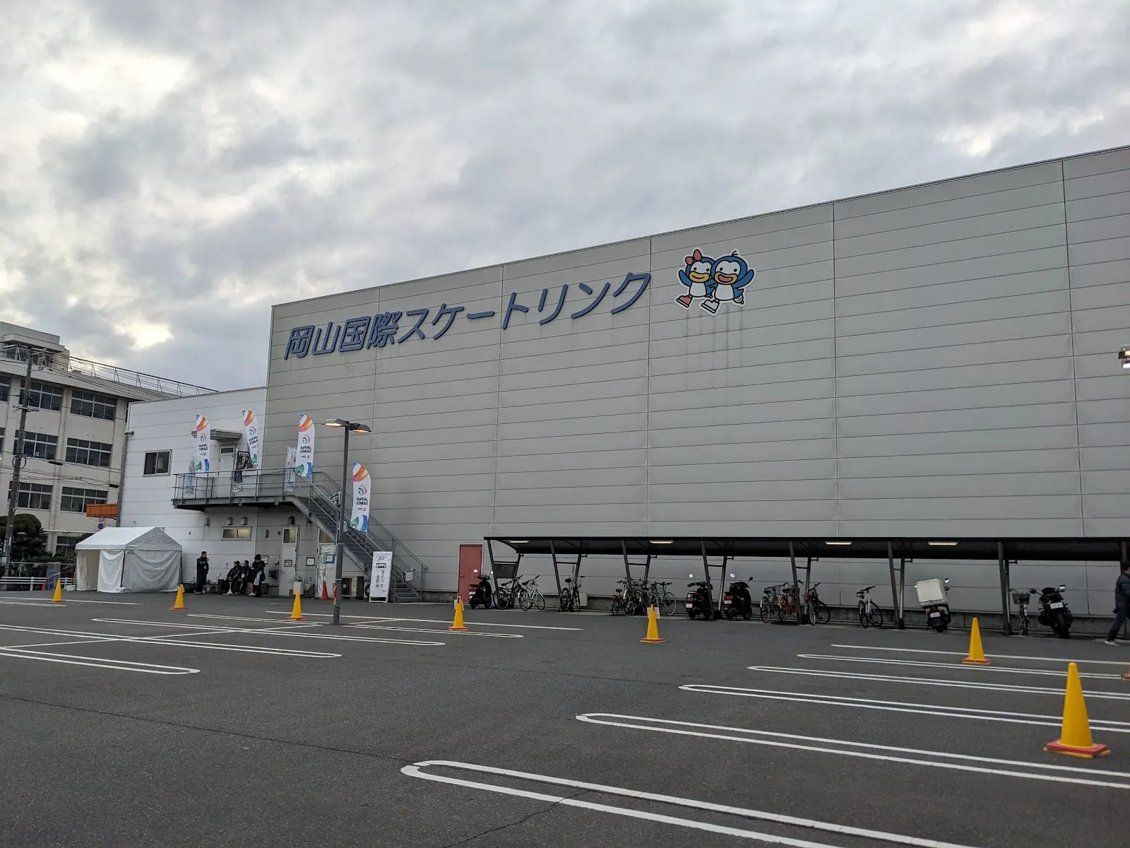 The grey front of the Okayama International Skating Rink seen from its car park, with the rink name and mascot across the wall — Okayama International Skating Rink