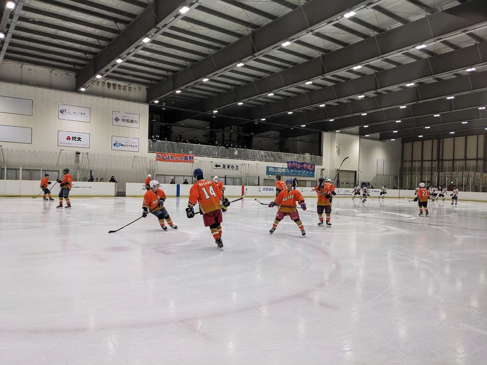 A game on the ice of the Okayama International Skating Rink, seen from the stands over the whole sheet under the black steel roof. — Okayama International Skating Rink