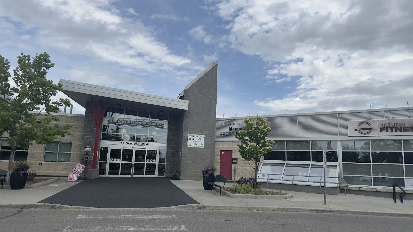 The Okotoks Recreation Centre entrance at 99 Okotoks Drive, with its name over the glass doors and the fitness centre wing to the right. — Okotoks Recreation Centre