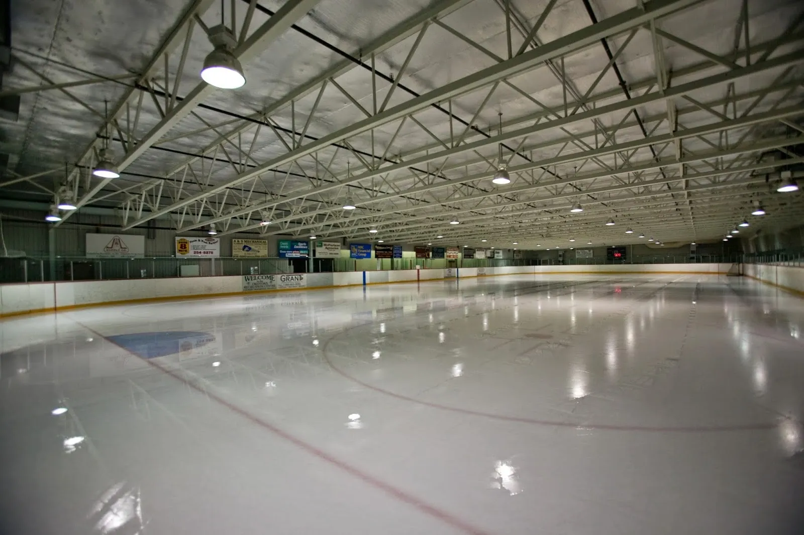 A wide empty sheet seen from the end, grey-blue ice with a faint circle under a long steel-trussed roof. — Okotoks Recreation Centre