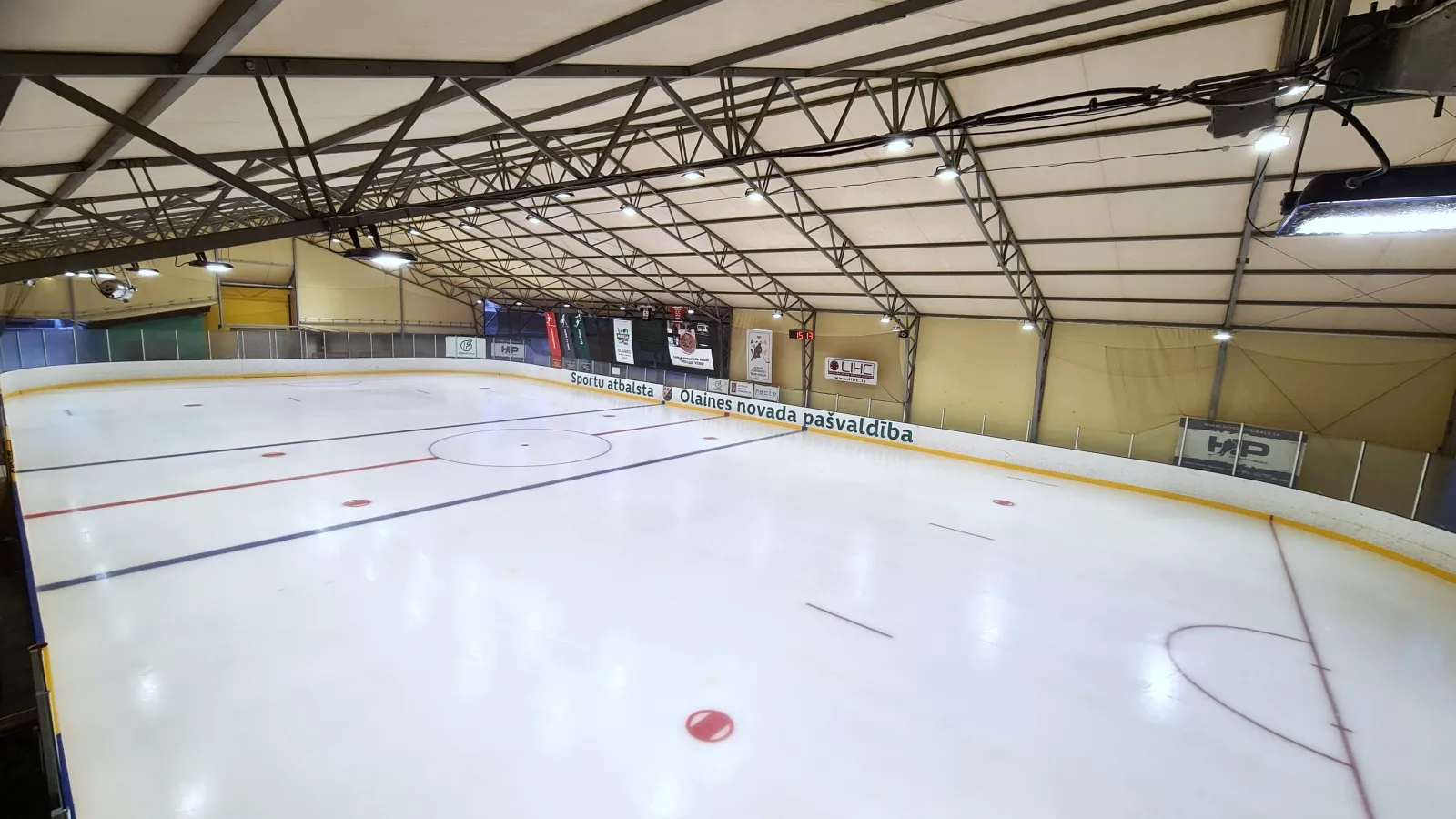 Olaines slidotavas ledus zem telts jumta, pie malām sponsoru reklāmas un Olaines novada pašvaldības nosaukums – Olaines slidotava