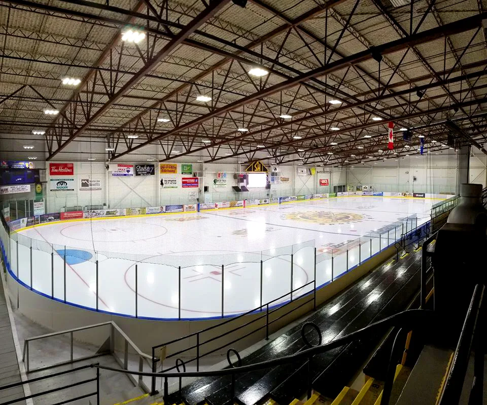 The main arena seen from the top of the stands: a full sheet under brown steel trusses, with sponsor signs on the far wall and a Canadian flag hanging from the roof. — Olds Sportsplex