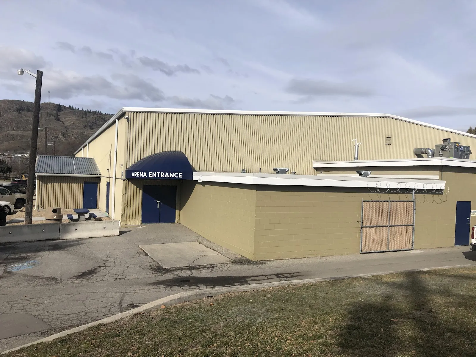 The tan metal-clad side of Oliver Arena with a navy awning reading Arena Entrance over a blue door, a wooden-clad service wall and a grassy verge, with hills behind under a pale sky. — Oliver & District Arena