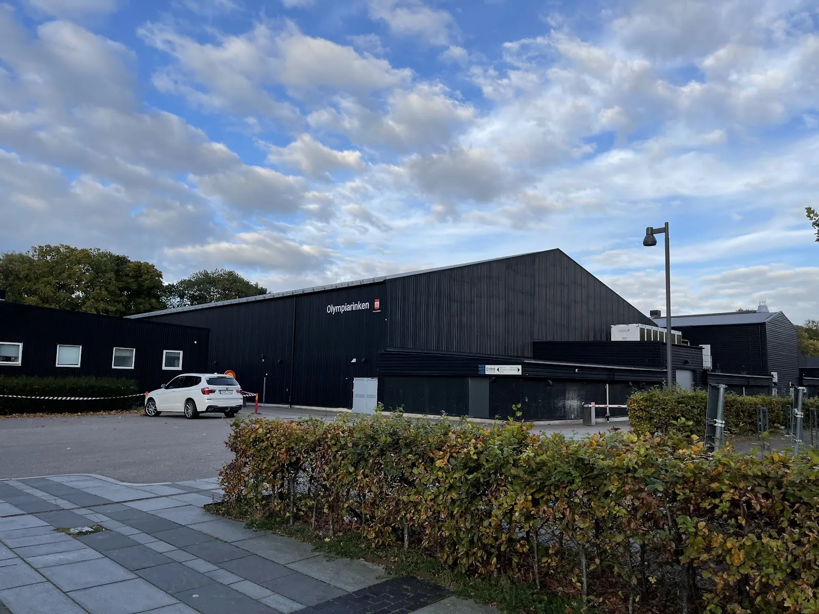 Den långa svartklädda Olympiarinken i Helsingborg med namnet på väggen, sedd över parkeringen under en blå himmel – Olympiarinken