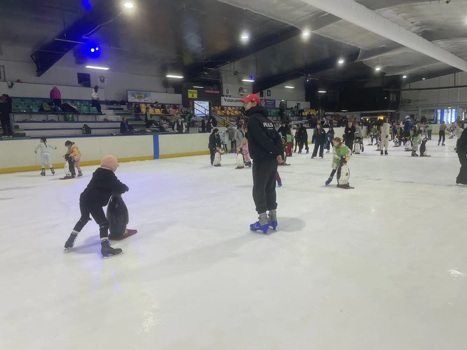 The wide view of the ice rink from the side: the whole sheet with public skaters at a distance and the boards and sponsor panels. — Olympic Ice Skating Centre