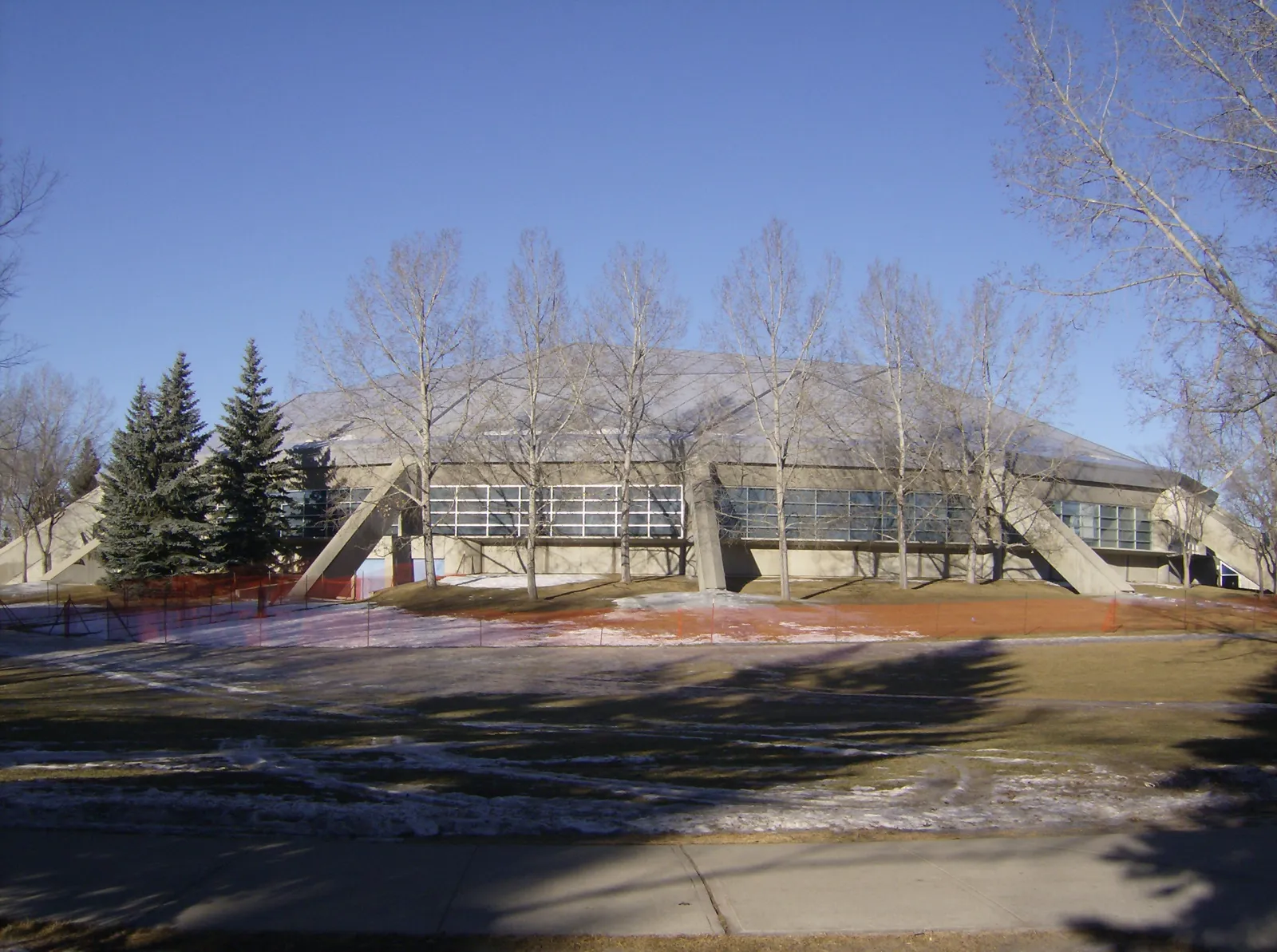 The Olympic Oval's long, low building with angled concrete buttresses and a band of windows, seen across a lawn through bare winter trees. — Olympic Oval