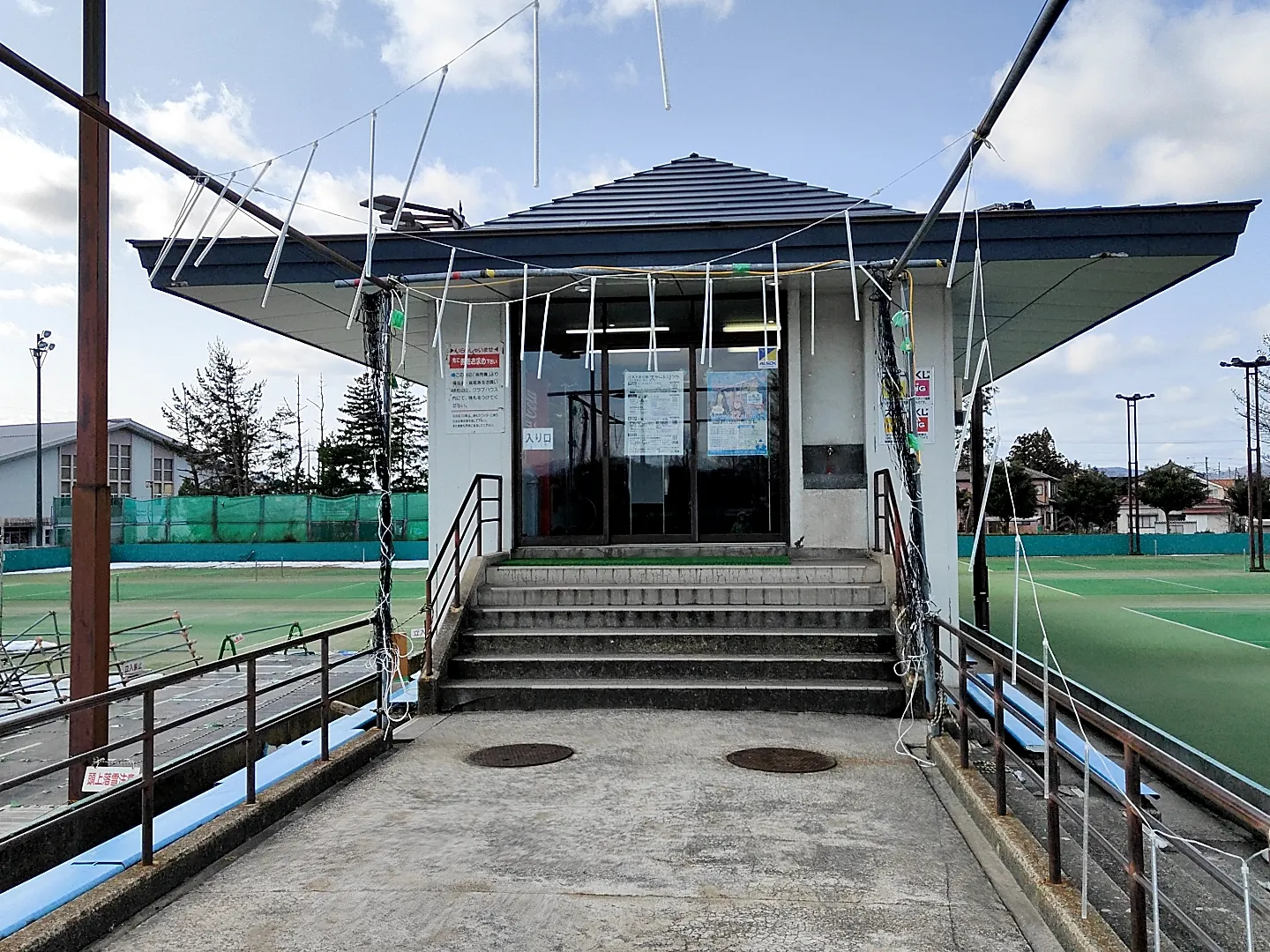 リンク脇の階段の上にある小さな入口の建物。背後に運動場が広がっています。 — Omagihara Skating Rink